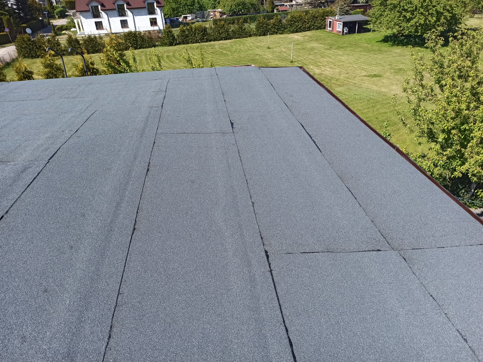 Widok z góry na płaski dach pokryty papą termozgrzewalną, z widocznymi łączeniami i teksturą, na tle zielonego trawnika i zabudowań.