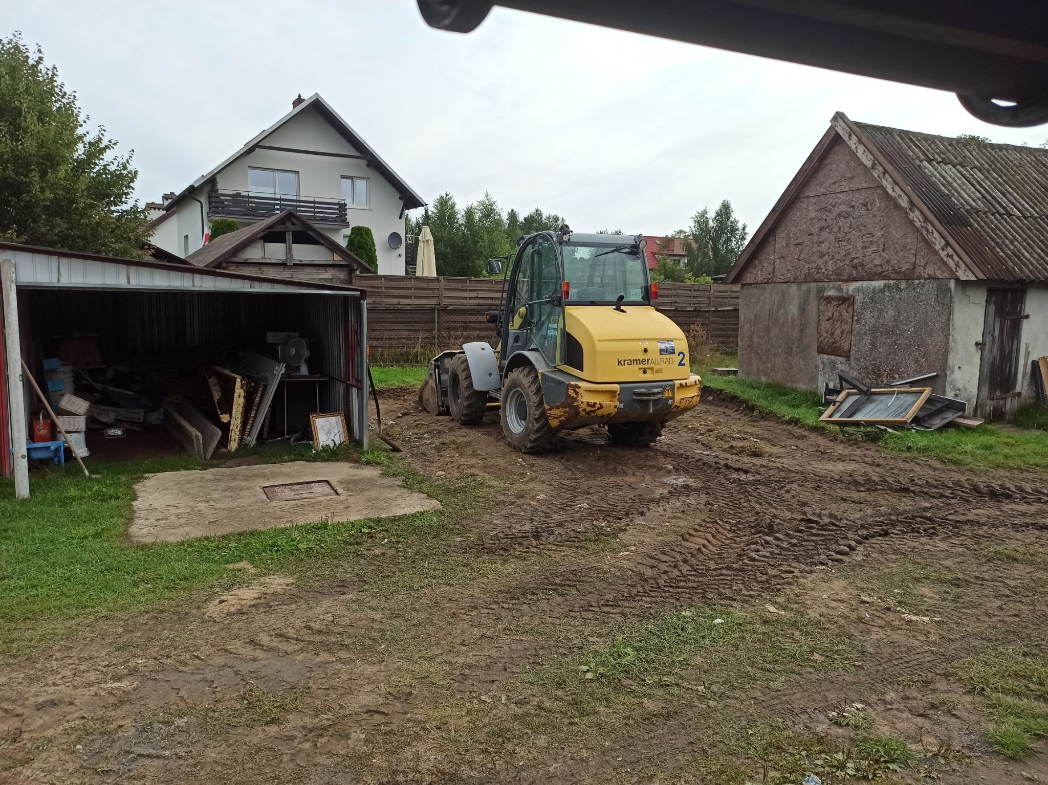 Żółta ładowarka Kramer Allrad 2 na błotnistym terenie z wyraźnymi śladami opon, garaż z otwartymi drzwiami po lewej stronie, dom jednorodzinny w tle.