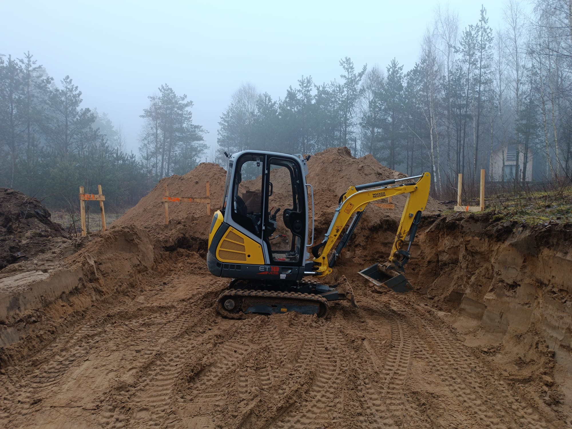Żółto-szara mini koparka Wacker Neuson ET18 na dnie wykopu pod fundamenty, widoczne ślady gąsienic, drewniane szalunki i mglisty las w tle.