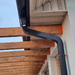 Zamontowany system rynnowy w kolorze czarnym, widoczny z boku budynku z drewnianą pergolą i elewacją wykończoną panelami drewnopodobnymi.