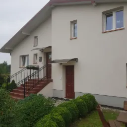 Jasny, dwukondygnacyjny dom z beżową elewacją, brązowymi drzwiami i schodami z czarną balustradą, widoczny z poziomu ogrodu z niskimi krzewami.