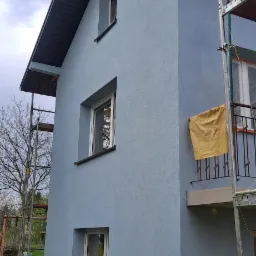 Świeżo otynkowana elewacja domu jednorodzinnego w odcieniach błękitu, widok z boku z fragmentem rusztowania i zabezpieczoną folią balkonem.