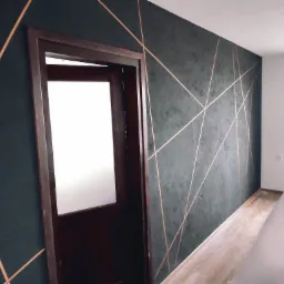 Ciemnoszara ściana z dekoracyjnymi, złotymi liniami tworzącymi geometryczny wzór, widoczna częściowo otwarta, ciemna rama drzwi i zabezpieczona folią podłoga.