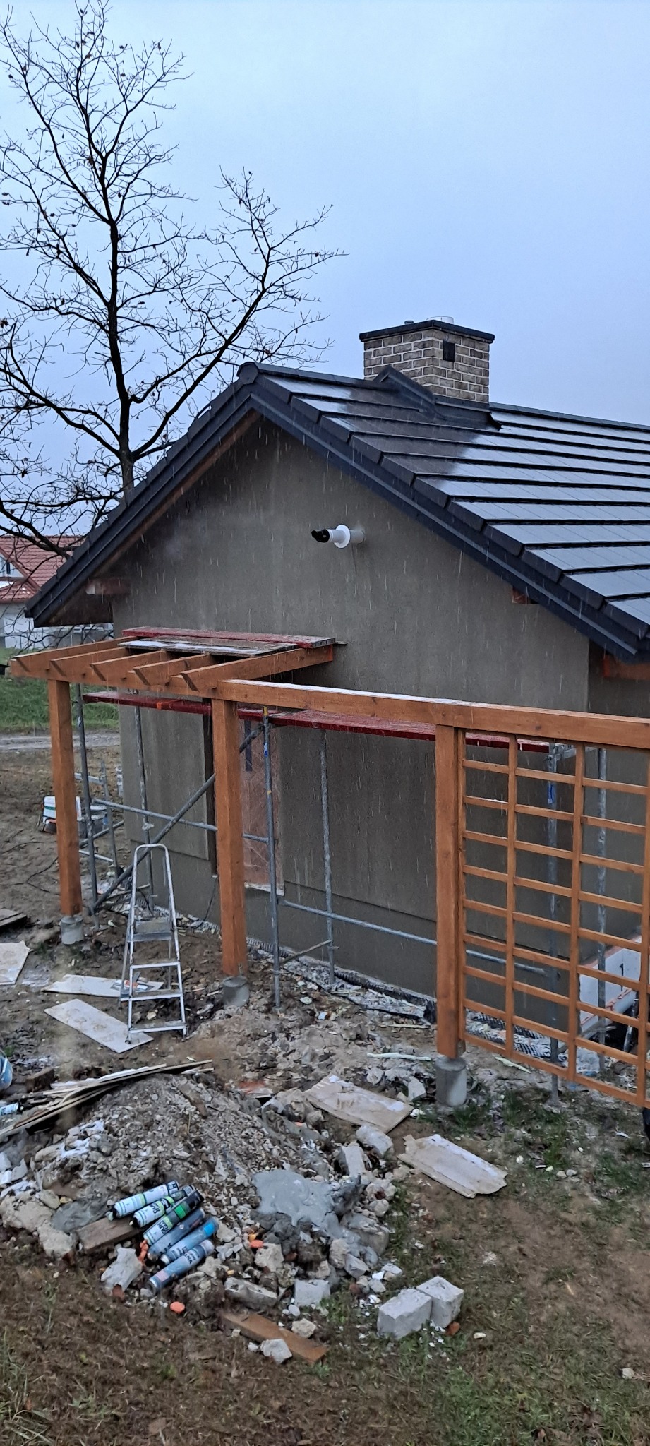 Dom w trakcie prac elewacyjnych, widoczne rusztowania, drewniana konstrukcja pergoli, świeżo otynkowana ściana i resztki materiałów budowlanych na ziemi w deszczowy dzień.