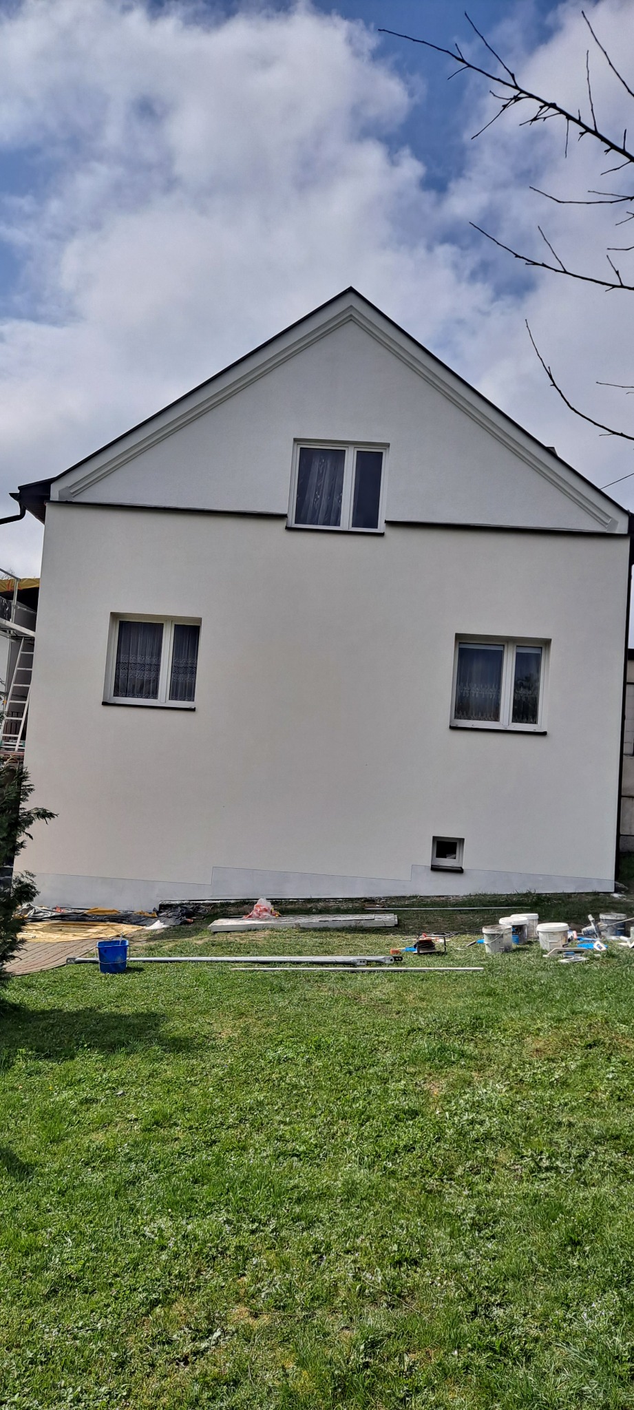 Dwukondygnacyjny dom z białymi ścianami po remoncie, widoczne okna z białymi ramami i ciemnymi zasłonami, na trawniku leżą narzędzia i materiały budowlane, w tle pochmurne niebo.