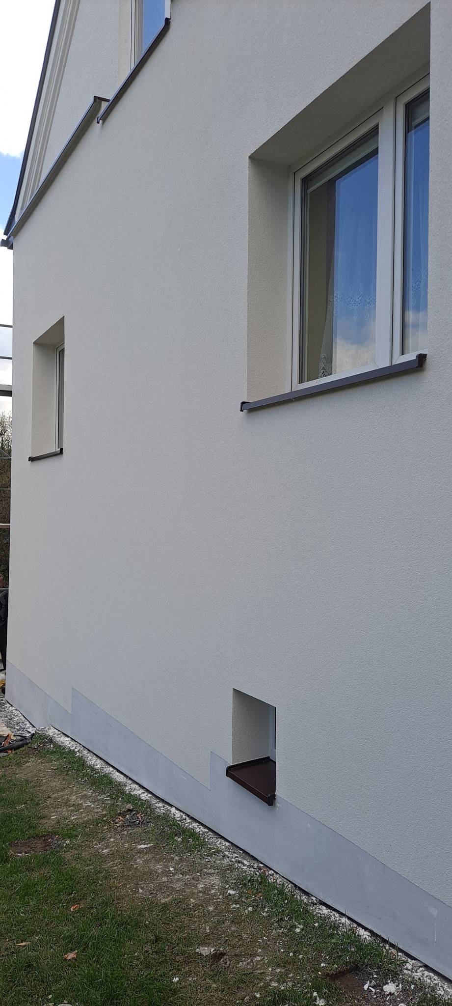 Świeżo otynkowana, jasna elewacja budynku z widocznymi oknami i fragmentem trawnika u podstawy ściany. Dolna część elewacji pomalowana na szaro, oddzielona od reszty. Małe okienko wentylacyjne...