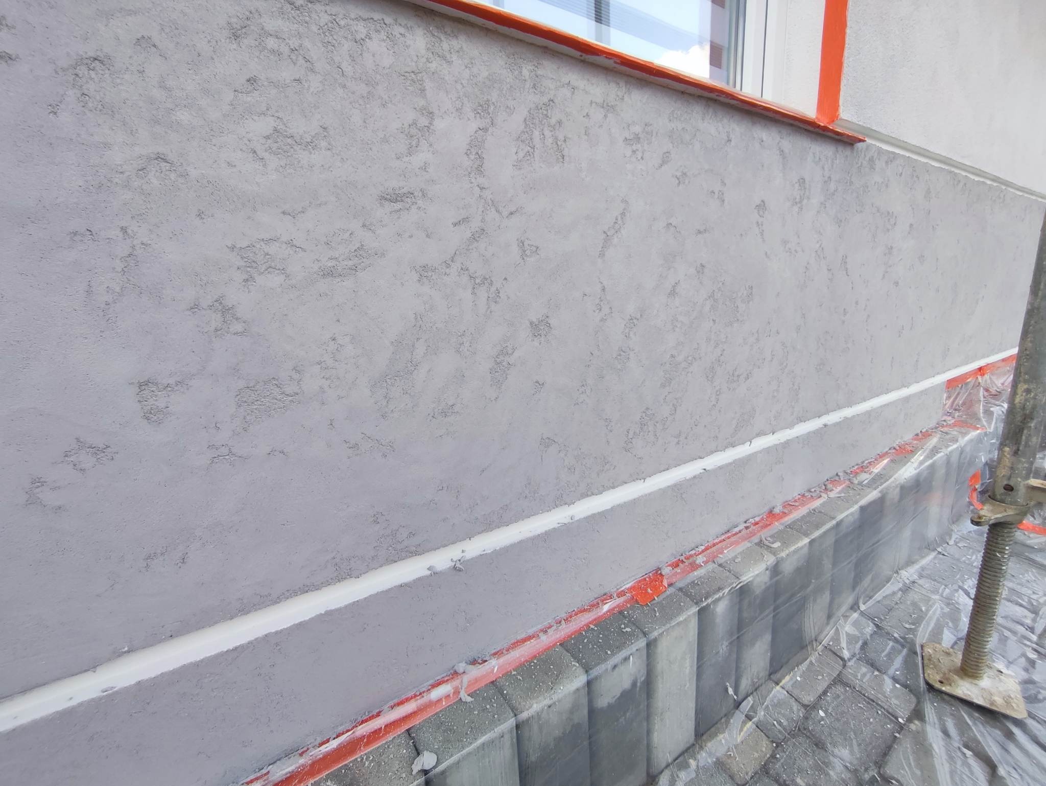 Świeżo położony szary tynk strukturalny na elewacji domu, zabezpieczony folią i taśmą malarską, widoczny fragment rusztowania.