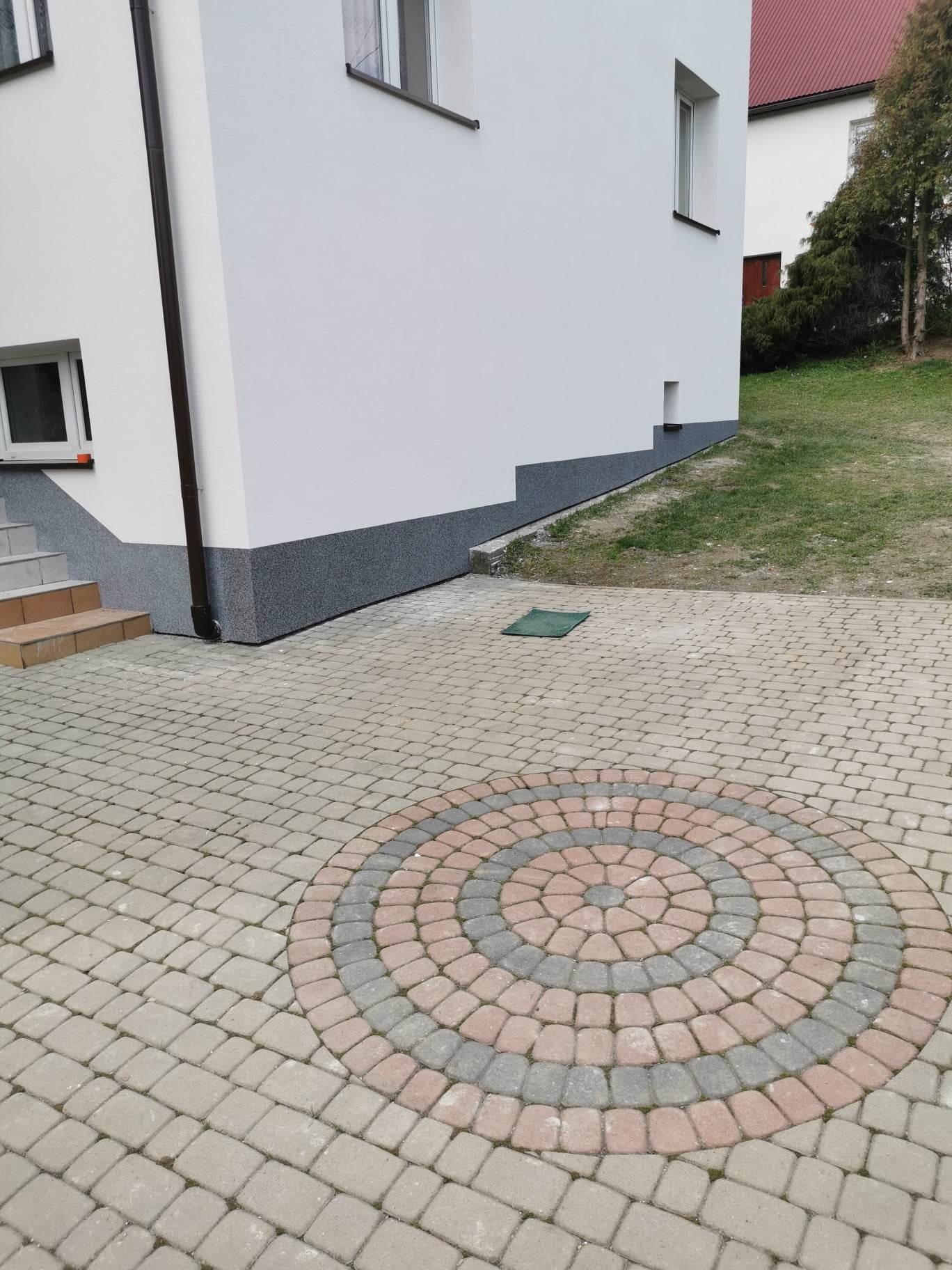 Wykończona elewacja budynku z widoczną strukturą tynku mozaikowego na cokole, białą ścianą i brązową rynną, na pierwszym planie kostka brukowa z dekoracyjnym wzorem koła.