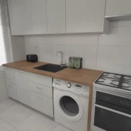 Jasna kuchnia z zabudową pod oknem, drewnianym blatem, zlewem, płytą grzewczą, piekarnikiem i pralką pod blatem.