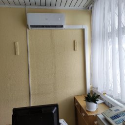 Reku-Vent Wentylacja, Klimatyzacja Pompy Ciepła - Biały klimatyzator AUX zamontowany na żółtej ścianie w biurze, z widocznymi białymi listwami maskującymi, biurkiem z monitorem i zasłoniętym oknem.
