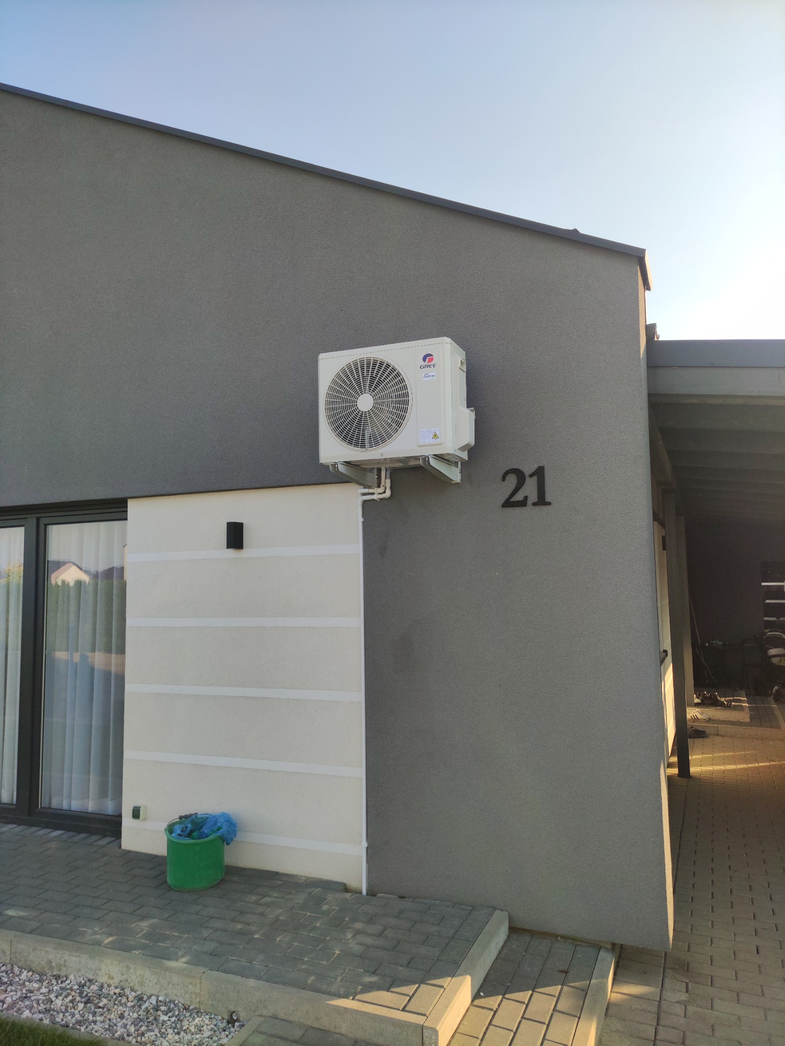 Zewnętrzna jednostka klimatyzacji marki Gree zamontowana na szarej elewacji budynku z numerem 21, obok okna z zasłonami i zielonego wiadra.