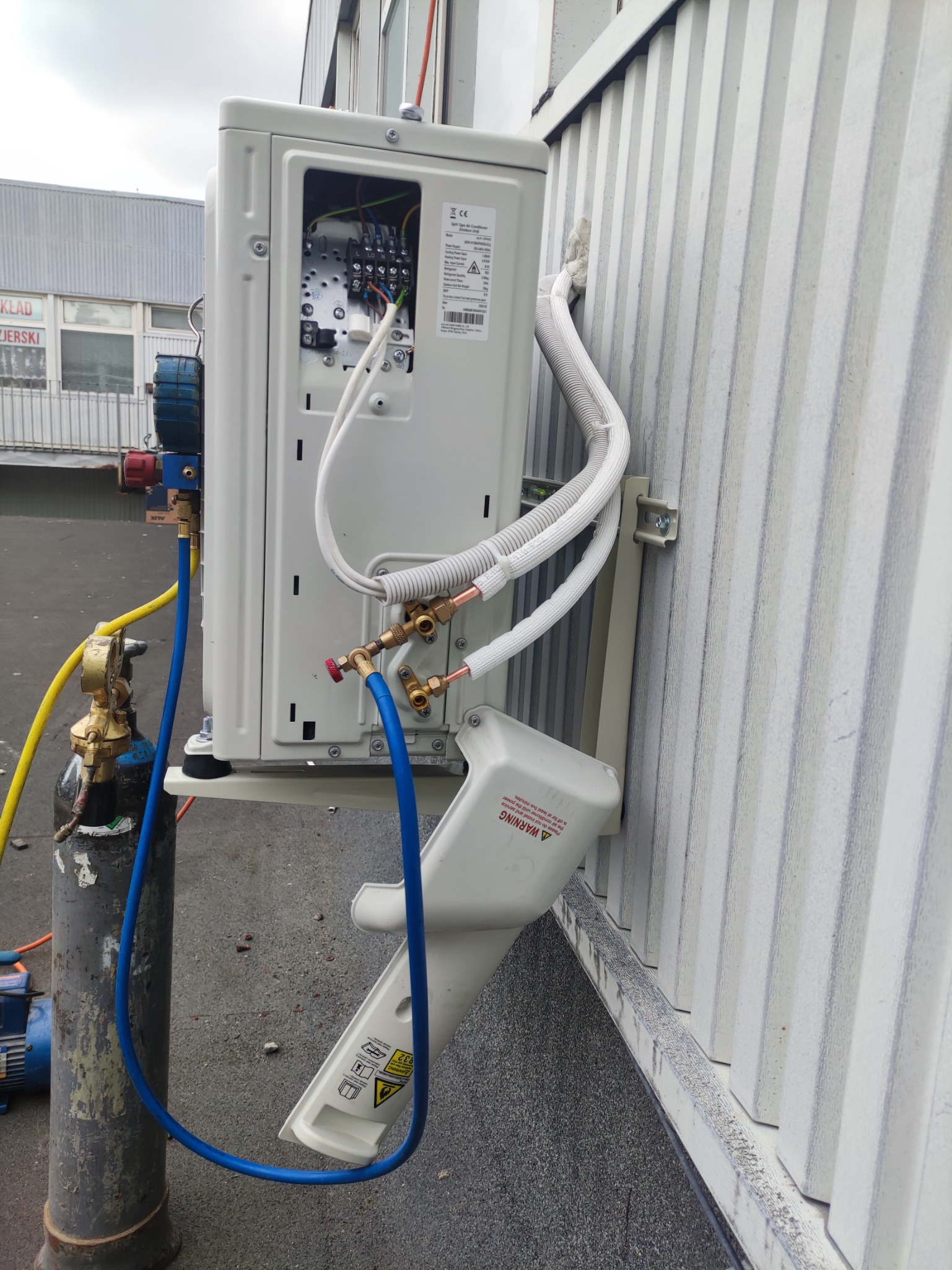 Zewnętrzna jednostka klimatyzacji zamontowana na ścianie budynku, podłączona do butli z czynnikiem chłodniczym podczas serwisu, widoczne przewody i złącza. Otwarta obudowa ukazuje wewnętrzne...