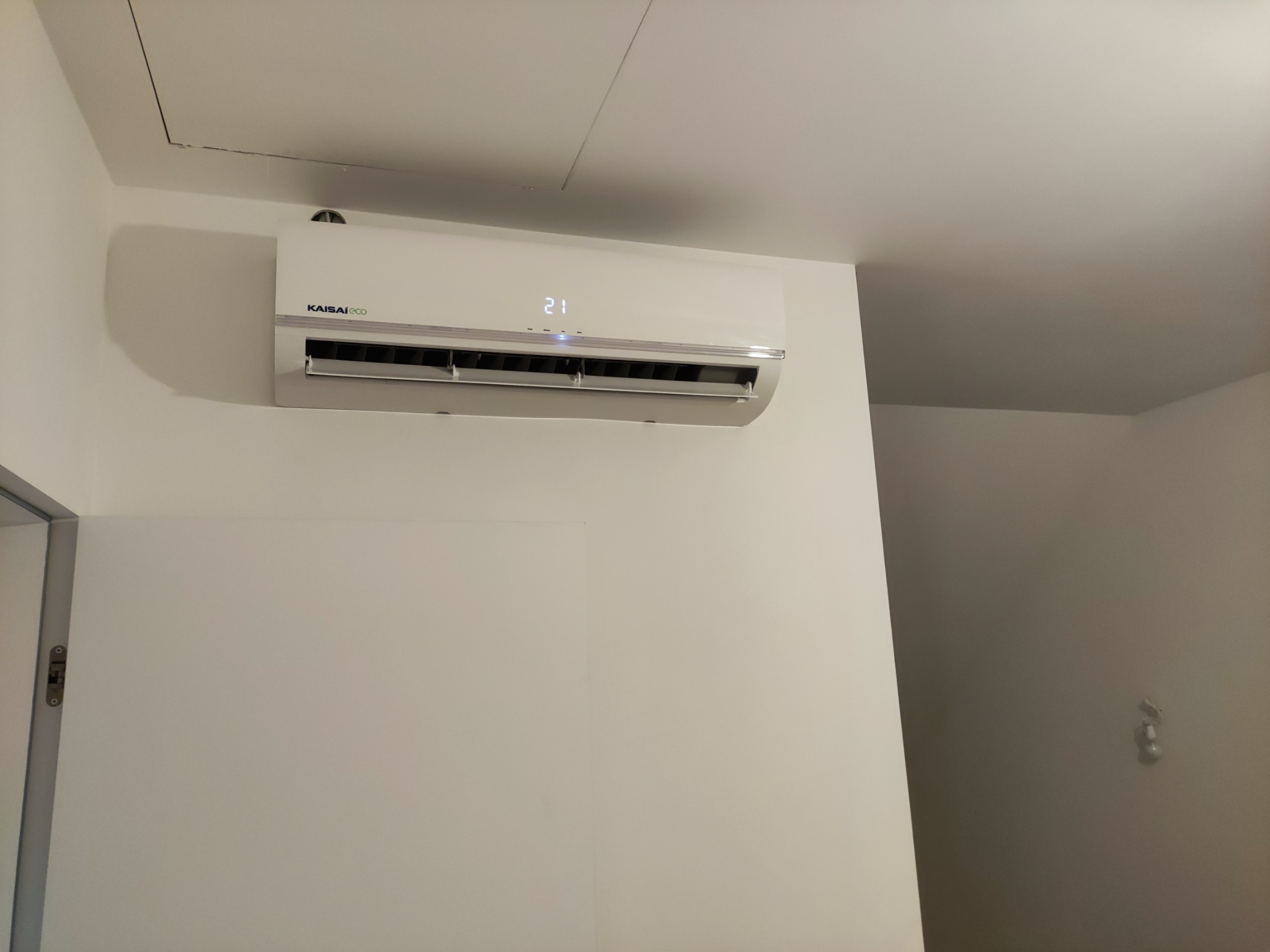 Biała jednostka klimatyzacyjna Kaisaieco zamontowana na ścianie w pomieszczeniu, wyświetlająca temperaturę 21 stopni, widoczny fragment sufitu z klapą rewizyjną.