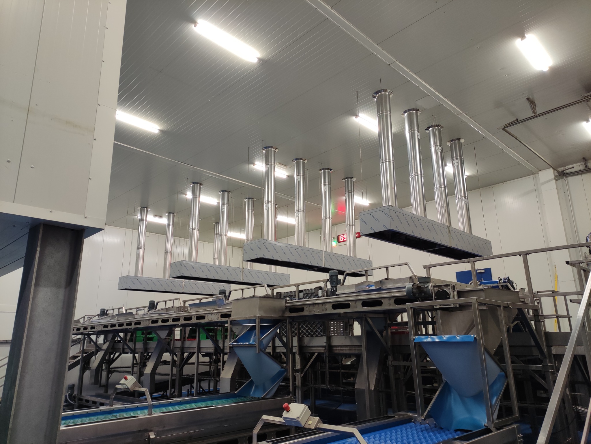 System wentylacji przemysłowej z rzędami rur wyciągowych w hali produkcyjnej oświetlonej lampami LED, widoczne elementy maszyn i konstrukcji stalowych.