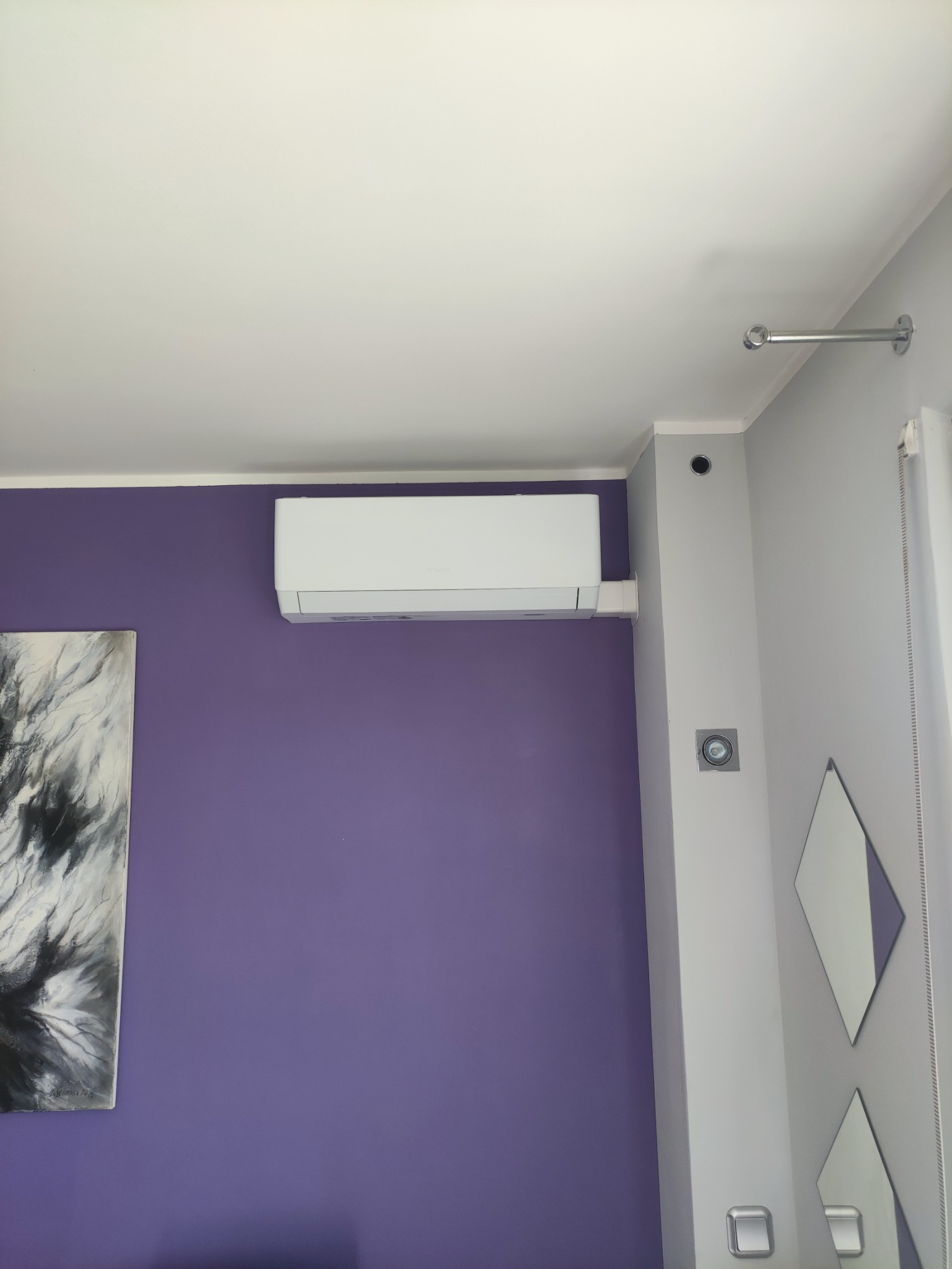 Biały klimatyzator zamontowany na fioletowej ścianie w pokoju z obrazem i lustrami, widok z dołu.
