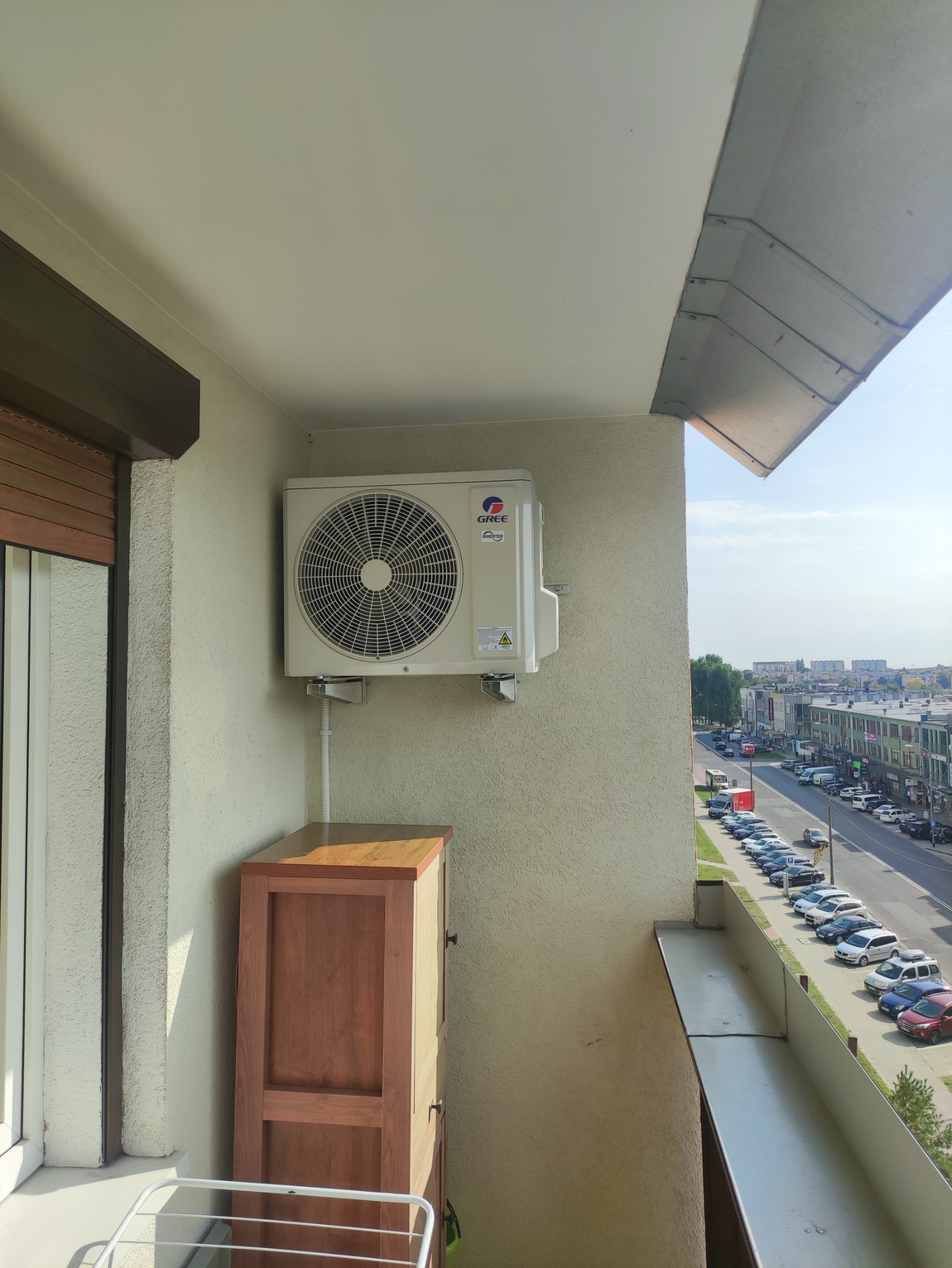 Zewnętrzna jednostka klimatyzacji marki Gree zamontowana na balkonie, widok na ulicę z zaparkowanymi samochodami i budynkami w tle, szafka na balkonie.