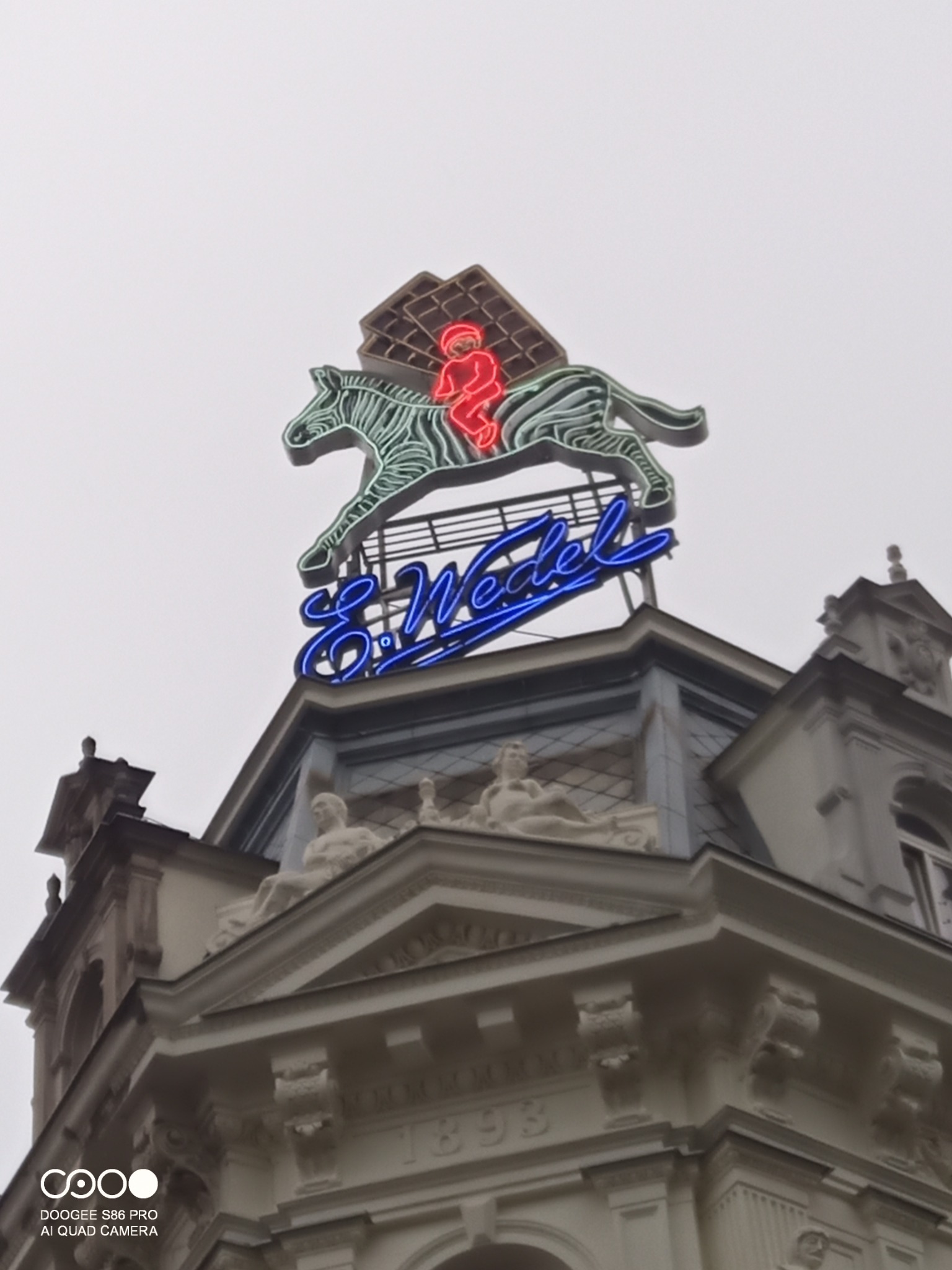 Neonowy szyld reklamowy E. Wedel z jeźdźcem na zebrze i tabliczką czekolady, umieszczony na dachu kamienicy z rzeźbami, widok z dołu.