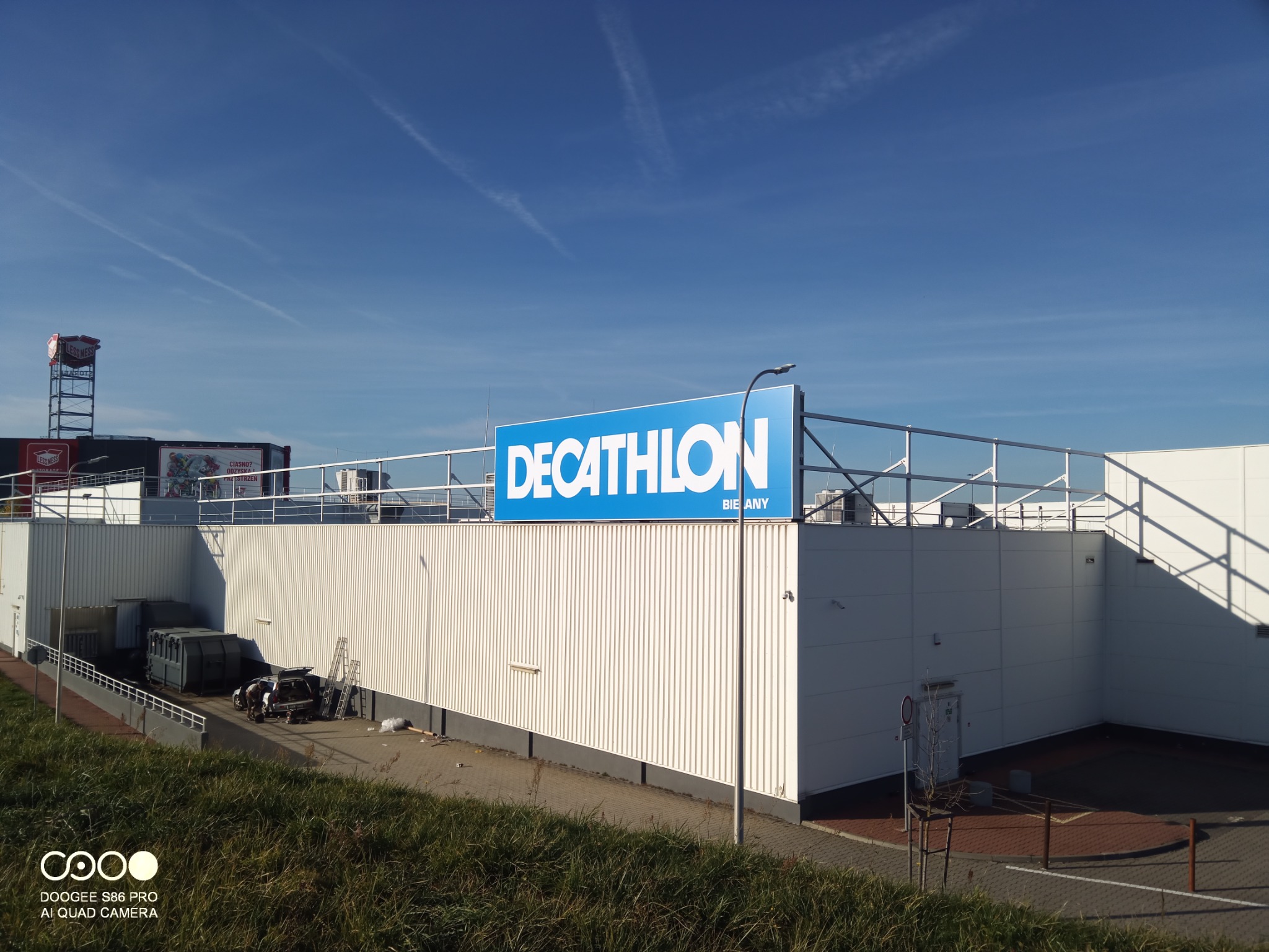 Budynek sklepu Decathlon z widocznym logo na dachu, z boku zaparkowany samochód z otwartym bagażnikiem i rozstawioną drabiną, sugerujący prace przy elewacji lub dachu.