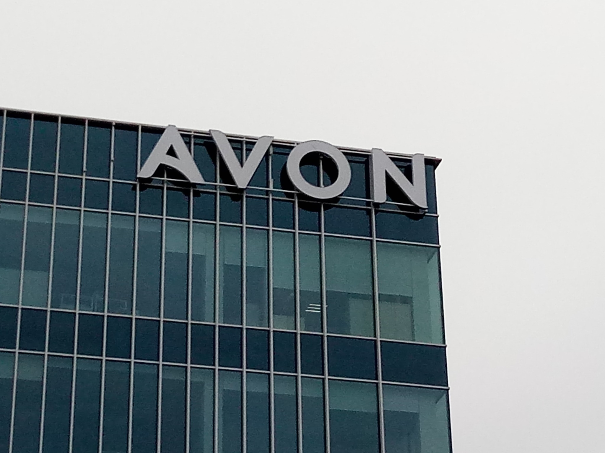 Metalowe litery AVON na szczycie nowoczesnego biurowca z ciemnymi szklanymi oknami pod pochmurnym niebem.