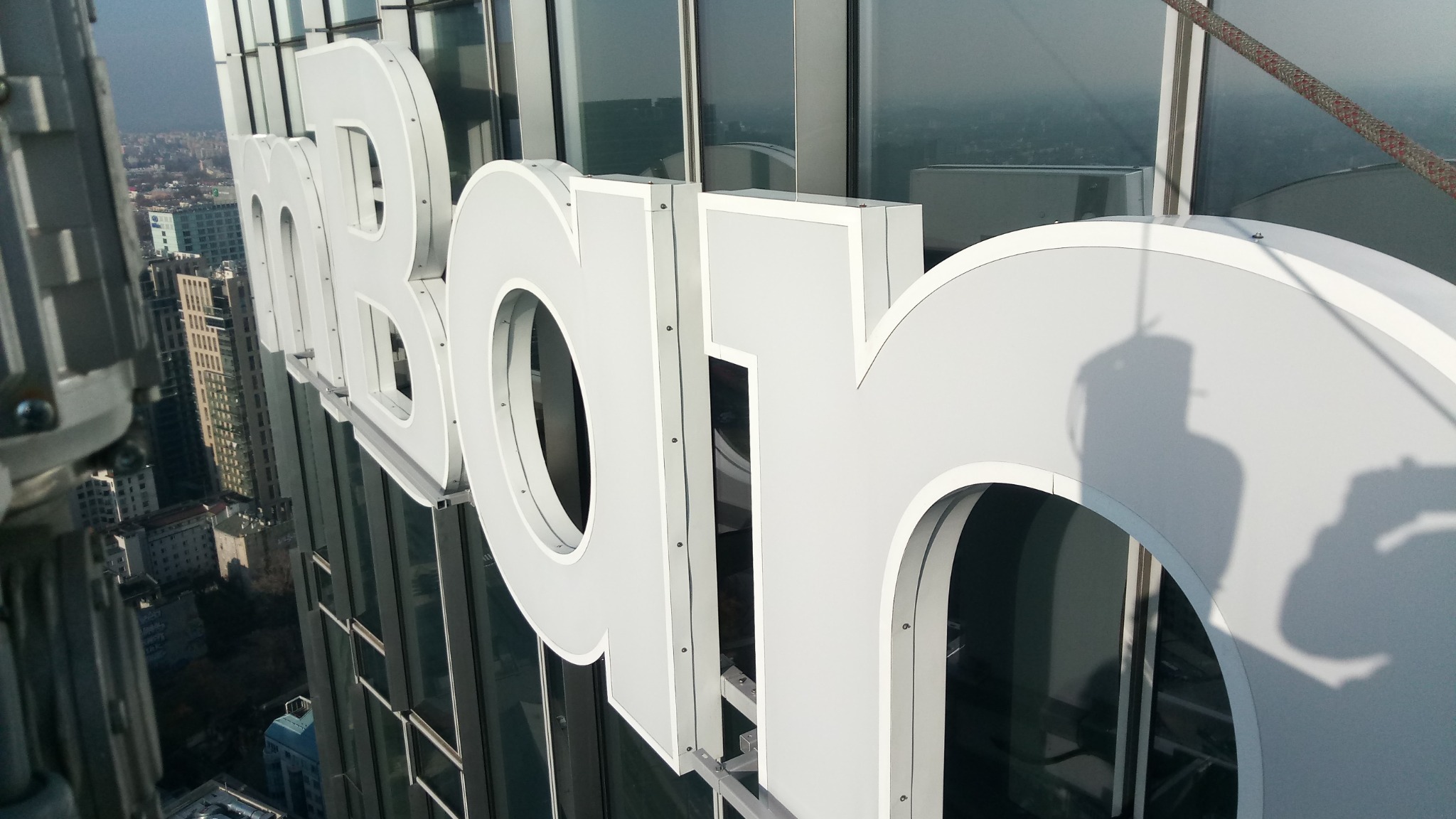 Montaż wielkoformatowych liter na elewacji wieżowca, widok z góry na miasto w tle, lina asekuracyjna widoczna w prawym górnym rogu.