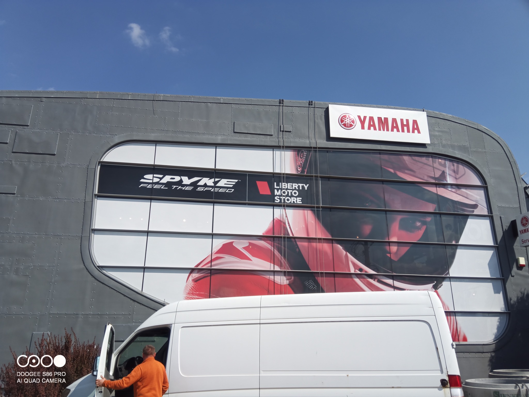 Szara elewacja budynku z dużymi reklamami Spyke, Liberty Moto Store i Yamaha, widoczny biały bus z otwartymi drzwiami i osoba w pomarańczowej bluzie.