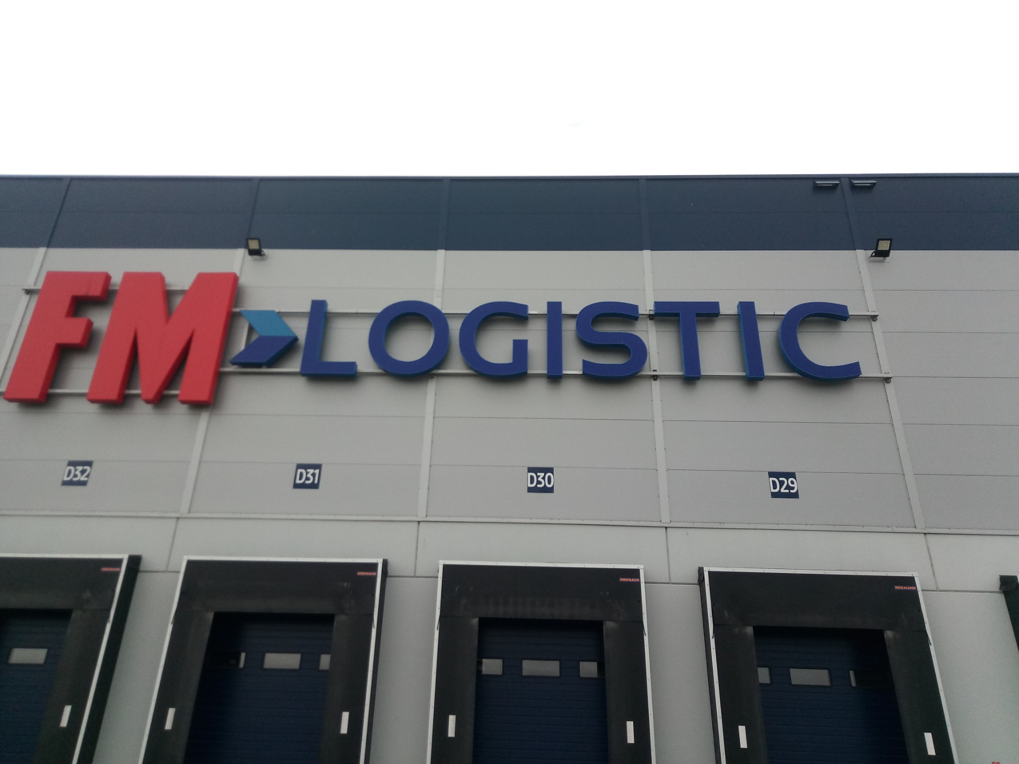 Elewacja budynku magazynowego z widocznym logo FM Logistic w kolorach czerwonym i niebieskim, nad czterema bramami dokowymi oznaczonymi numerami D32, D31, D30 i D29.