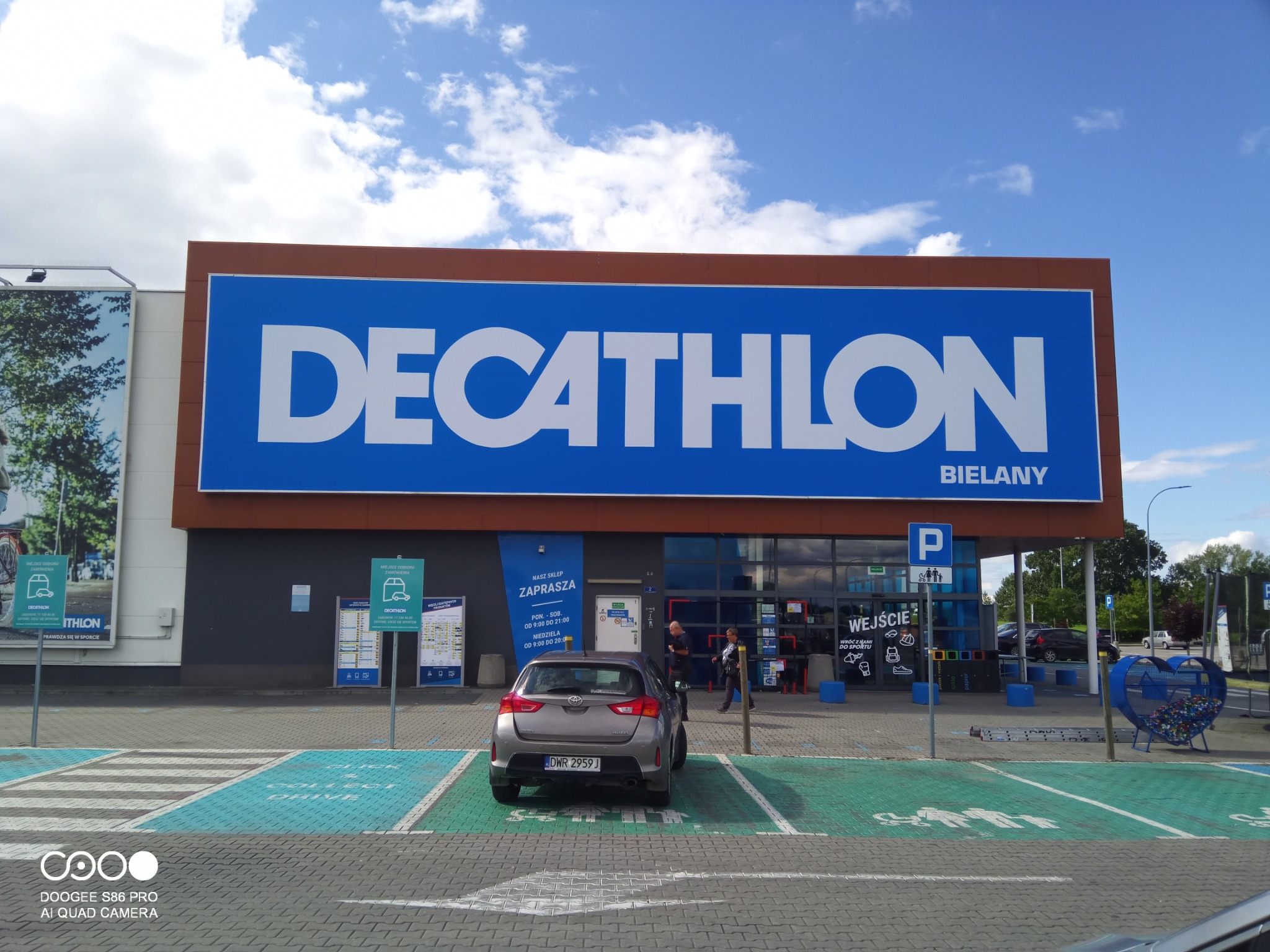 Duży, niebiesko-biały szyld sklepu sportowego Decathlon Bielany, umieszczony nad wejściem, widoczny z parkingu z zaparkowanym szarym samochodem osobowym.