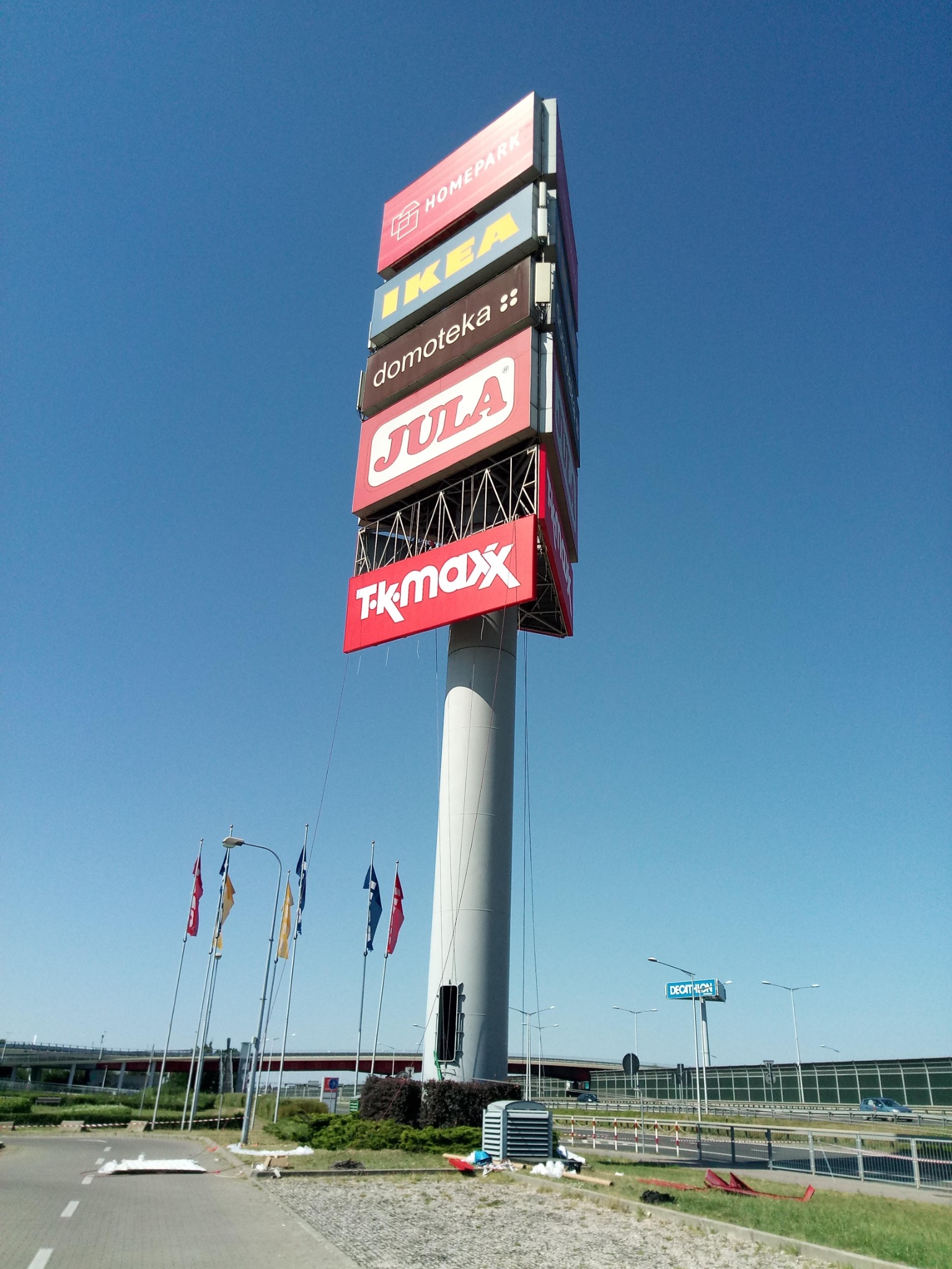 Wysoki pylon reklamowy z logotypami sklepów Homepark, IKEA, Domoteka, Jula i TK Maxx na tle błękitnego nieba, z widocznymi flagami i elementami infrastruktury drogowej w tle.
