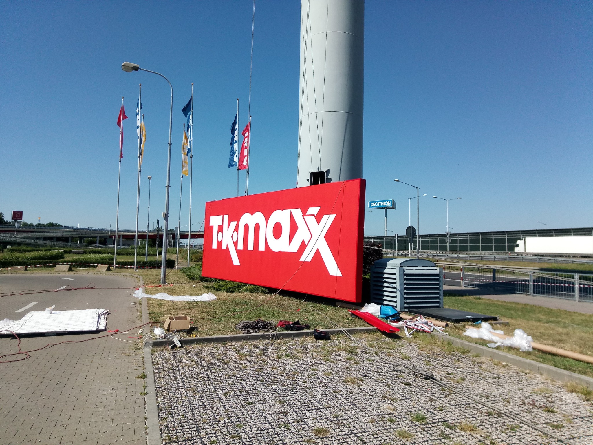 Montaż dużego, czerwonego szyldu TK Maxx z białym logo, widoczne narzędzia i materiały montażowe, wysoki słup w tle, błękitne niebo.