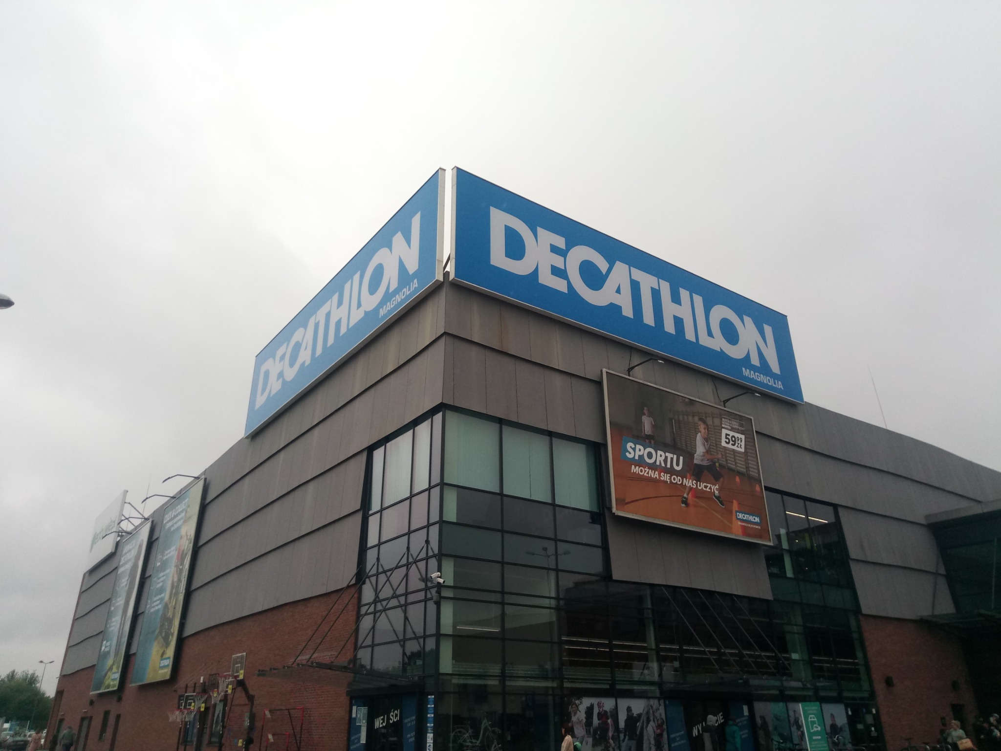 Budynek sklepu sportowego Decathlon Magnolia z dużym niebieskim szyldem na dachu i reklamami na elewacji, widok z dołu w pochmurny dzień.
