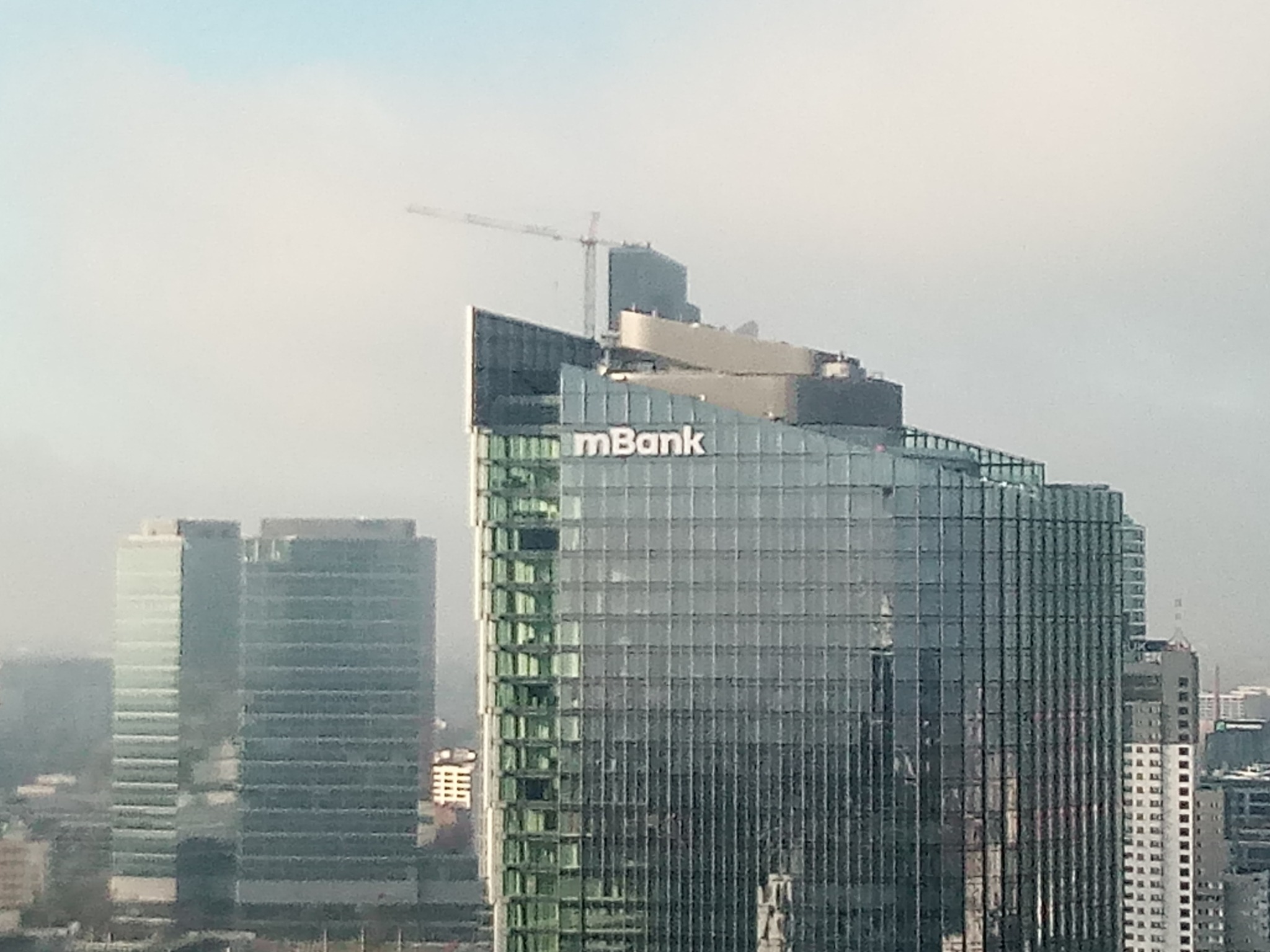 Wysoki szklany budynek biurowy z logo mBank na tle zamglonego miejskiego krajobrazu i żurawiem budowlanym na szczycie.