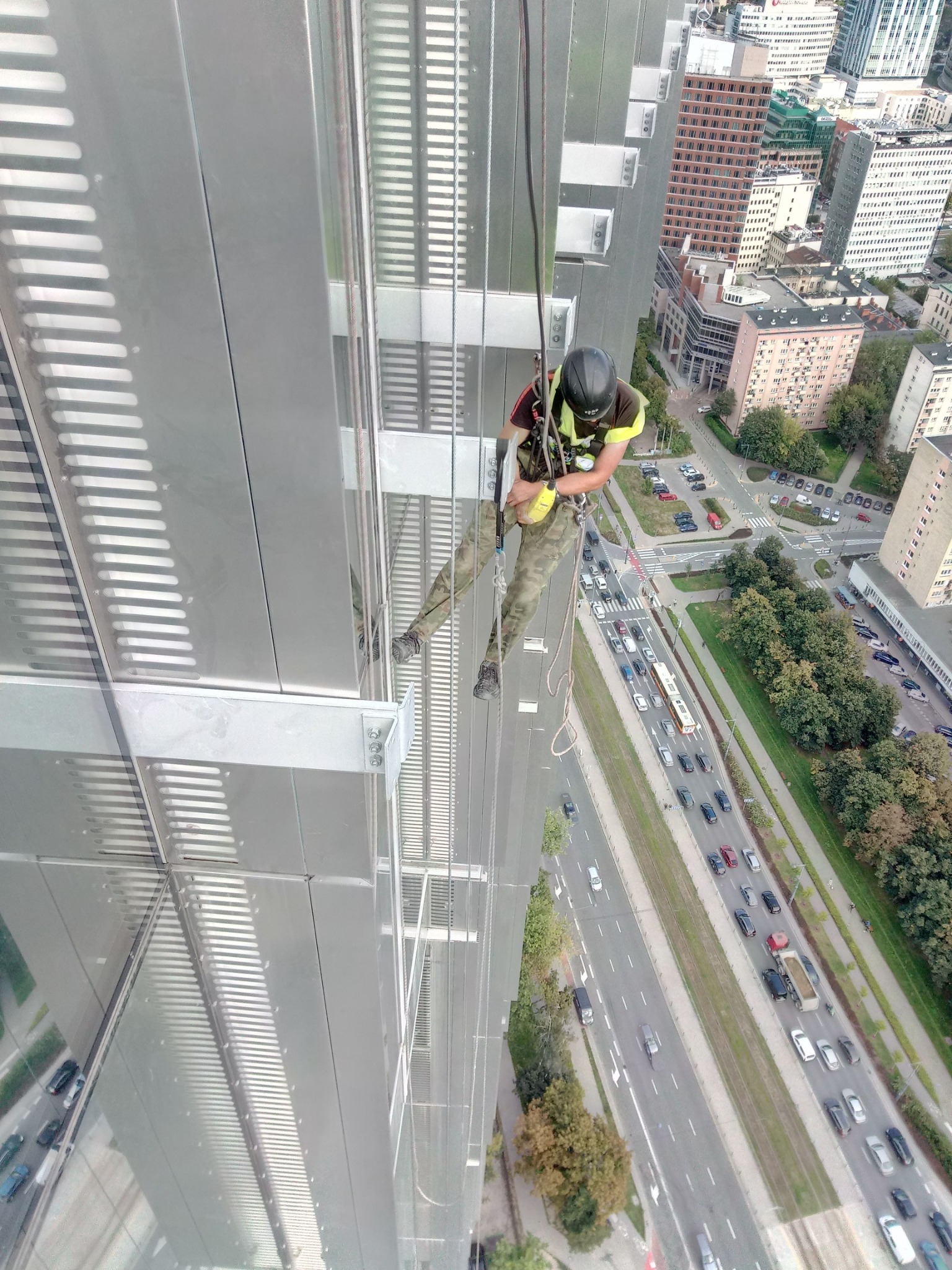 Pracownik w kasku i uprzęży, zawieszony na linie, wykonuje prace na wysokości na szklanej elewacji wieżowca z widokiem na ruchliwą ulicę miejską i zabudowę.