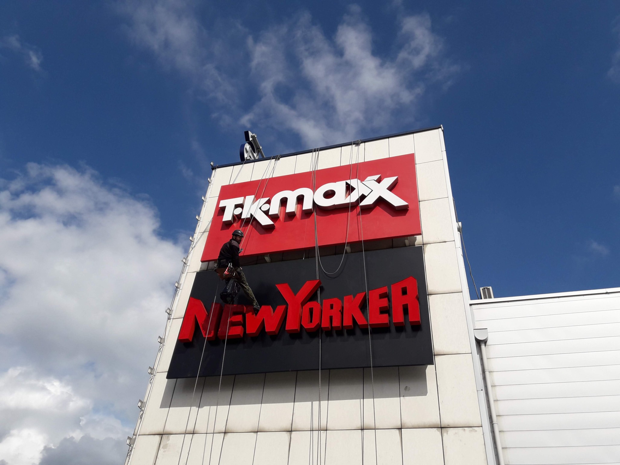 Dwóch alpinistów przemysłowych na linach przy elewacji budynku z dużymi szyldami TK Maxx i New Yorker, czyszczących lub konserwujących elewację i litery.