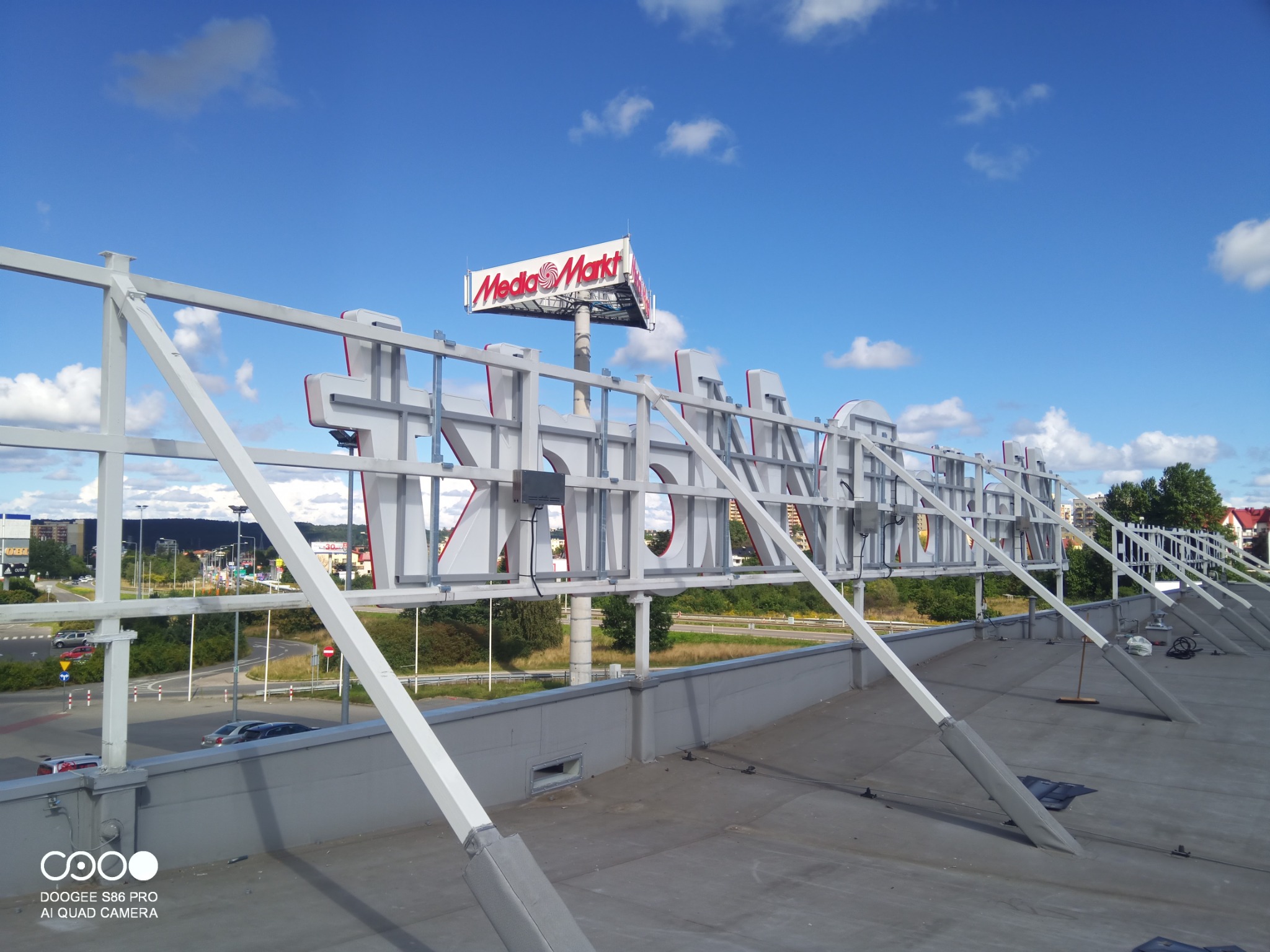 Montaż konstrukcji reklamowej Media Markt na dachu budynku w Ząbkach, widok z góry na białą stalową konstrukcję wsporczą i fragment krajobrazu miejskiego.