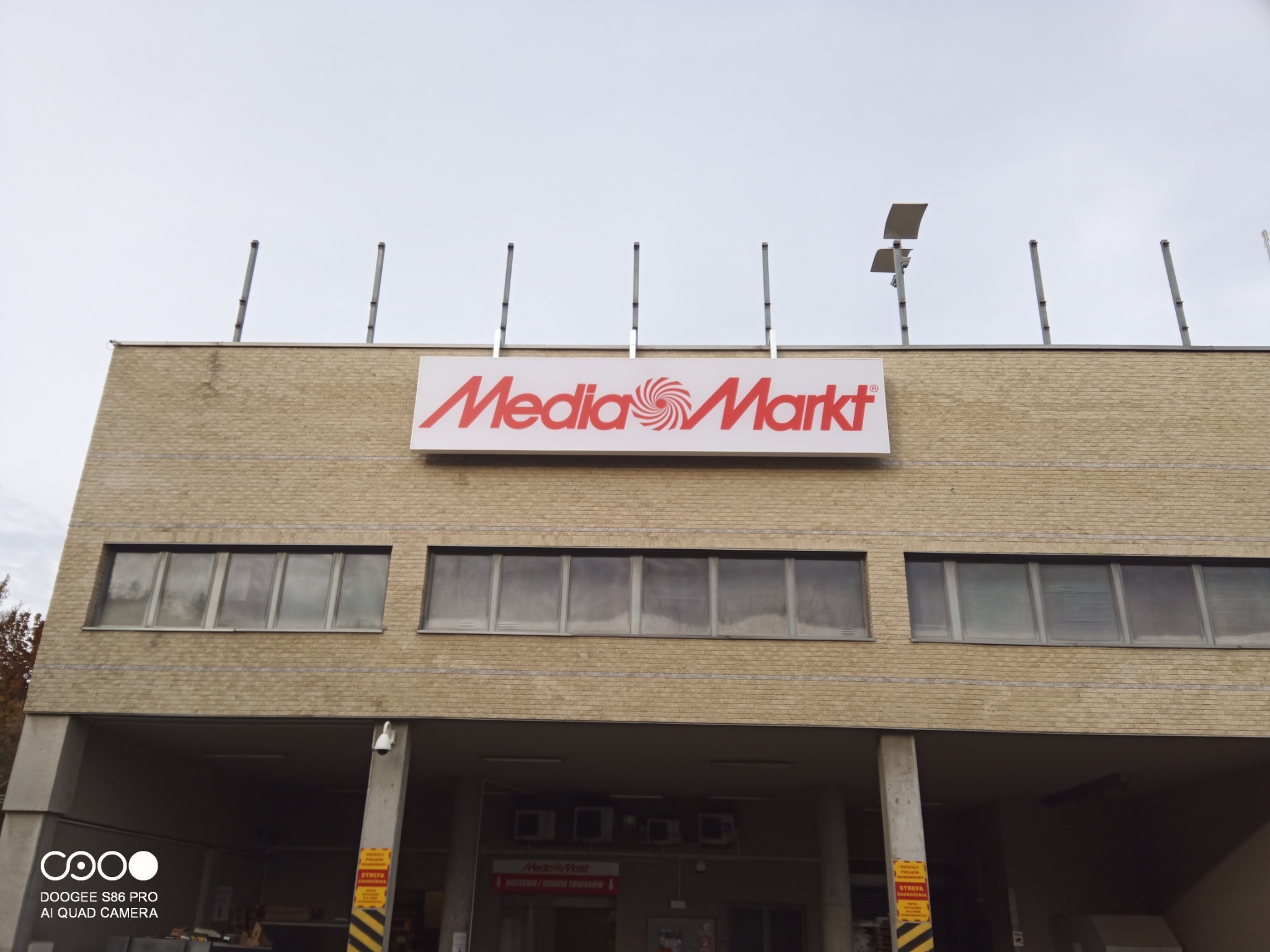 Budynek sklepu Media Markt z beżową ceglaną elewacją, widoczne okna i logo na dachu, z kilkoma antenami lub masztami na dachu.
