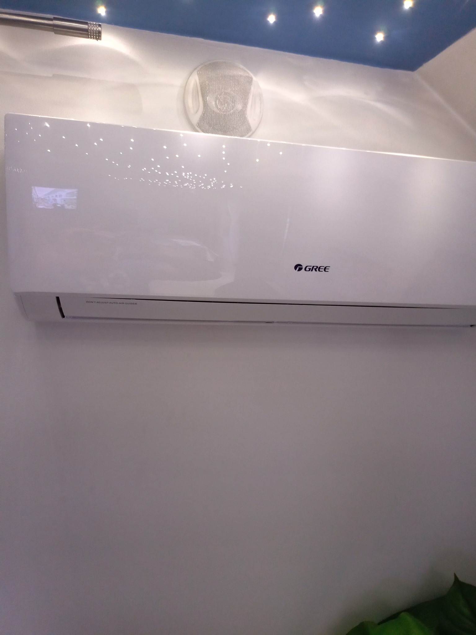 Biała jednostka wewnętrzna klimatyzatora marki Gree zamontowana na ścianie w pomieszczeniu z niebieskim sufitem i punktowym oświetleniem.