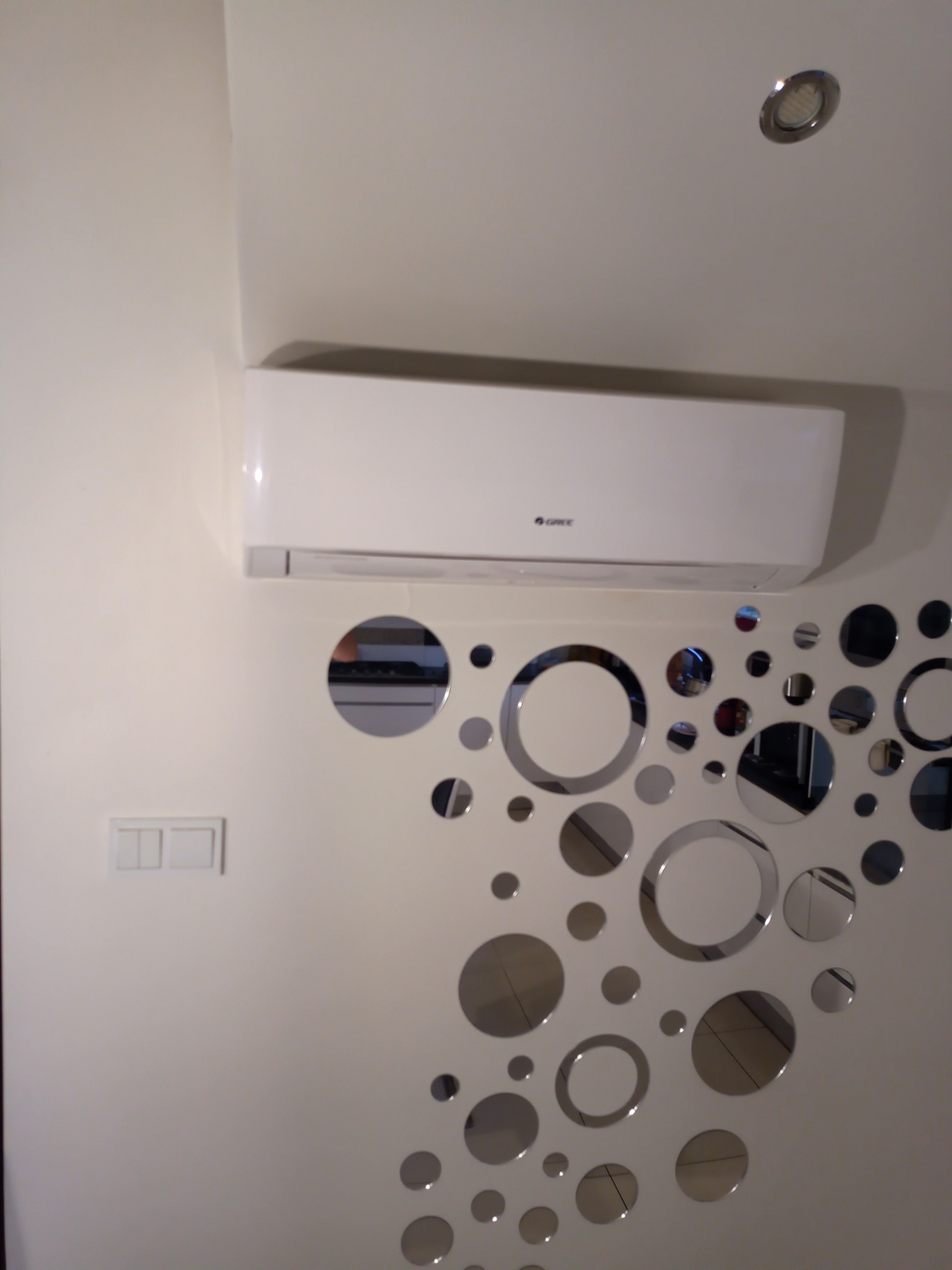 Biała jednostka klimatyzacji marki Gree, zamontowana na ścianie z dekoracyjnymi, okrągłymi lustrami w mieszkaniu w Kramsku, z widocznym włącznikiem światła.