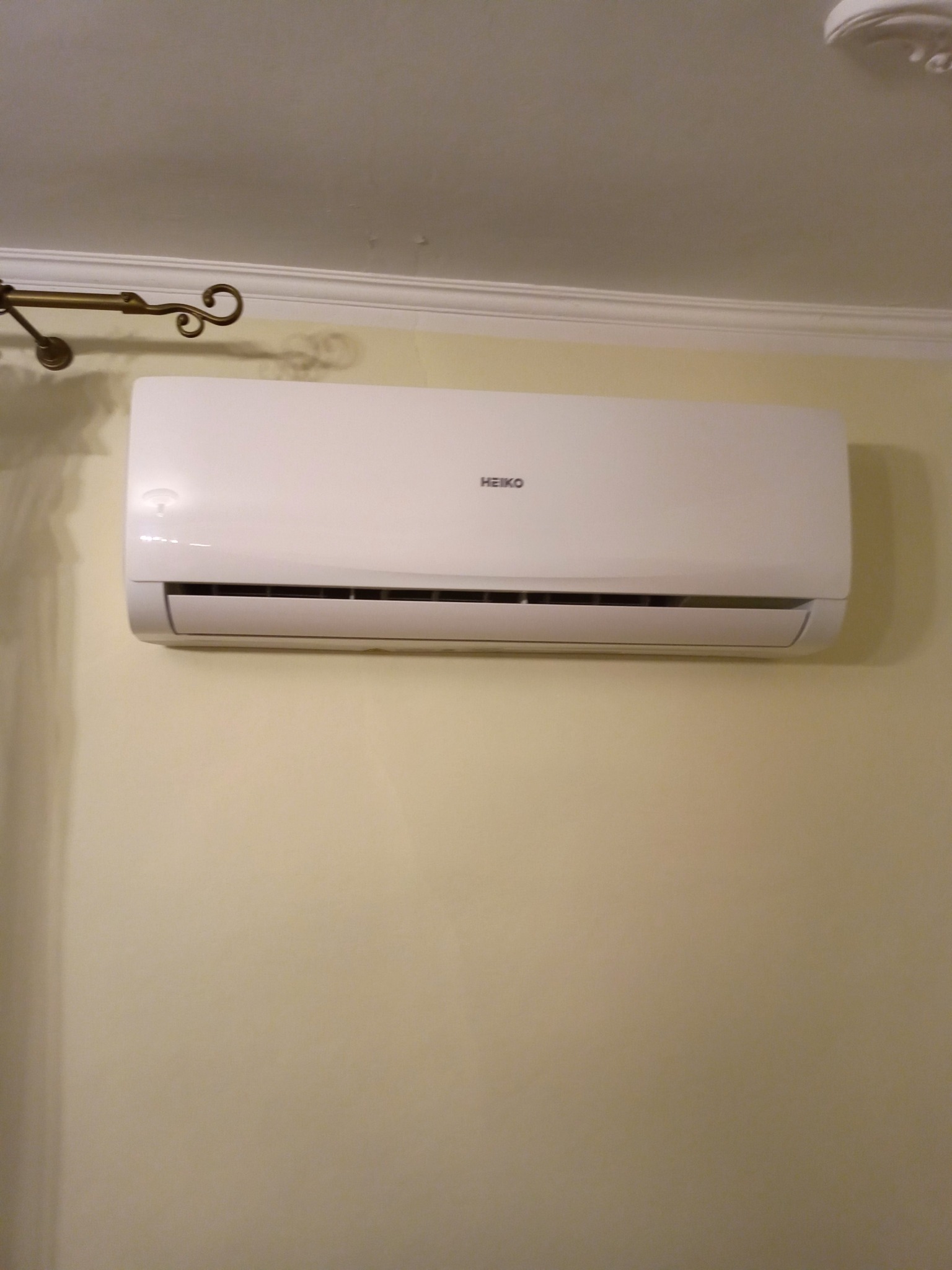 Biała jednostka klimatyzacyjna marki HEIKO zamontowana na kremowej ścianie w pomieszczeniu z ozdobnym gzymsem i fragmentem żyrandola.
