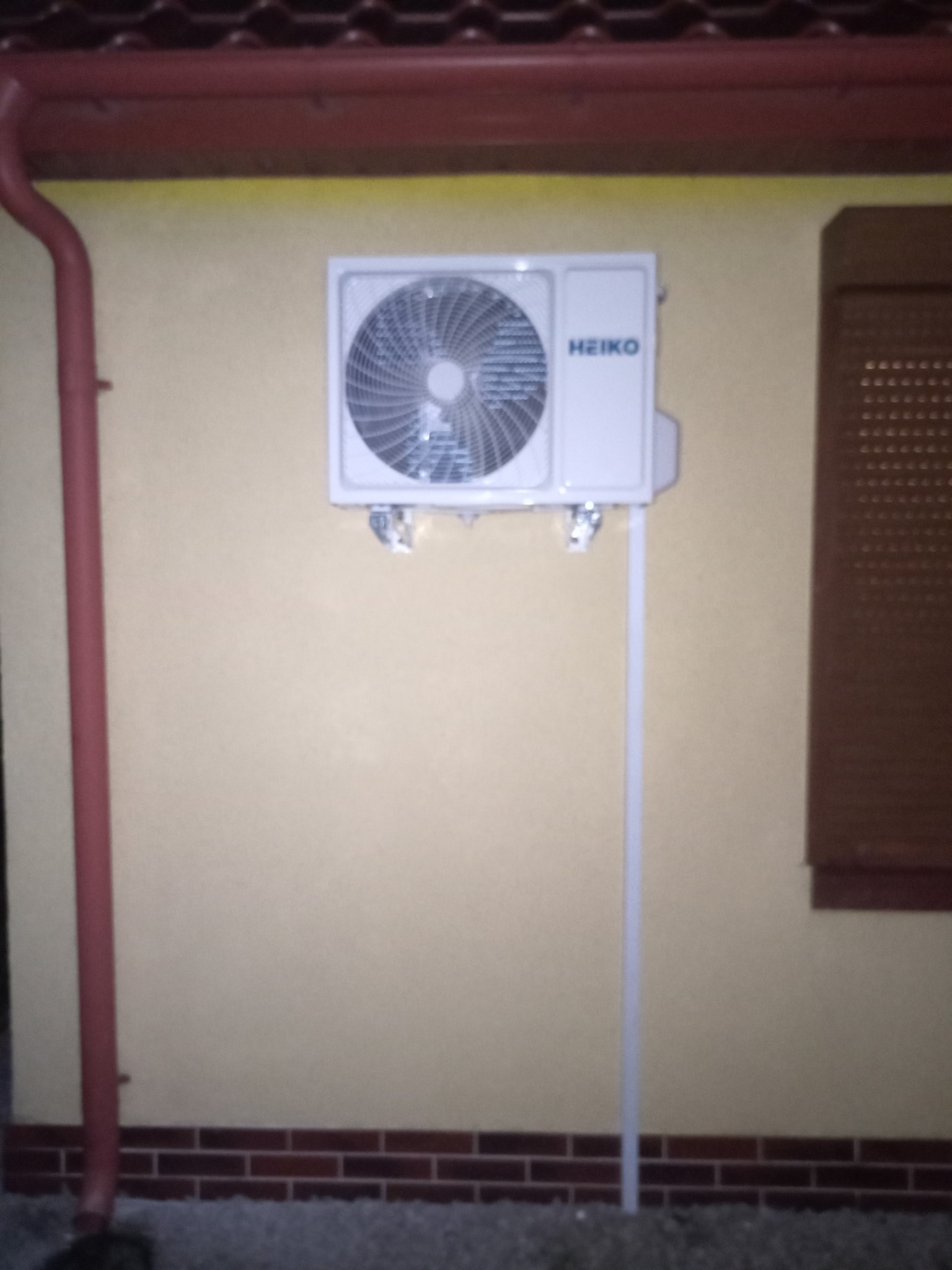 Zewnętrzna jednostka klimatyzatora marki Heiko zamontowana na elewacji domu z brązowymi roletami i rynną, wsparta na metalowej podporze.