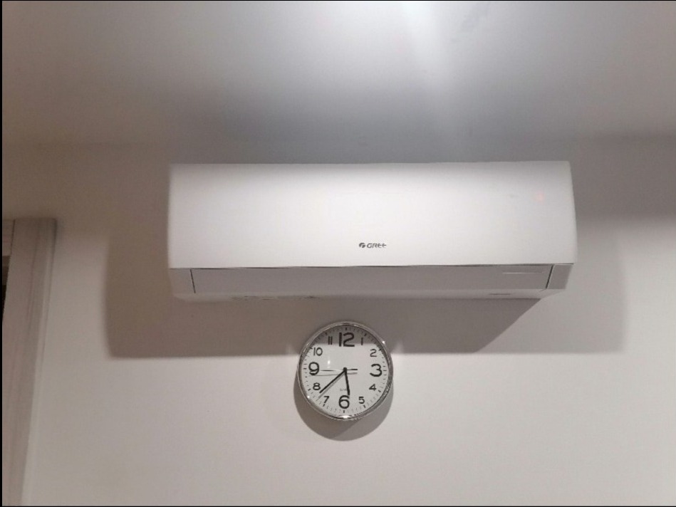 Biały klimatyzator marki Gree zawieszony na białej ścianie, pod nim okrągły zegar wskazujący godzinę 6:30.