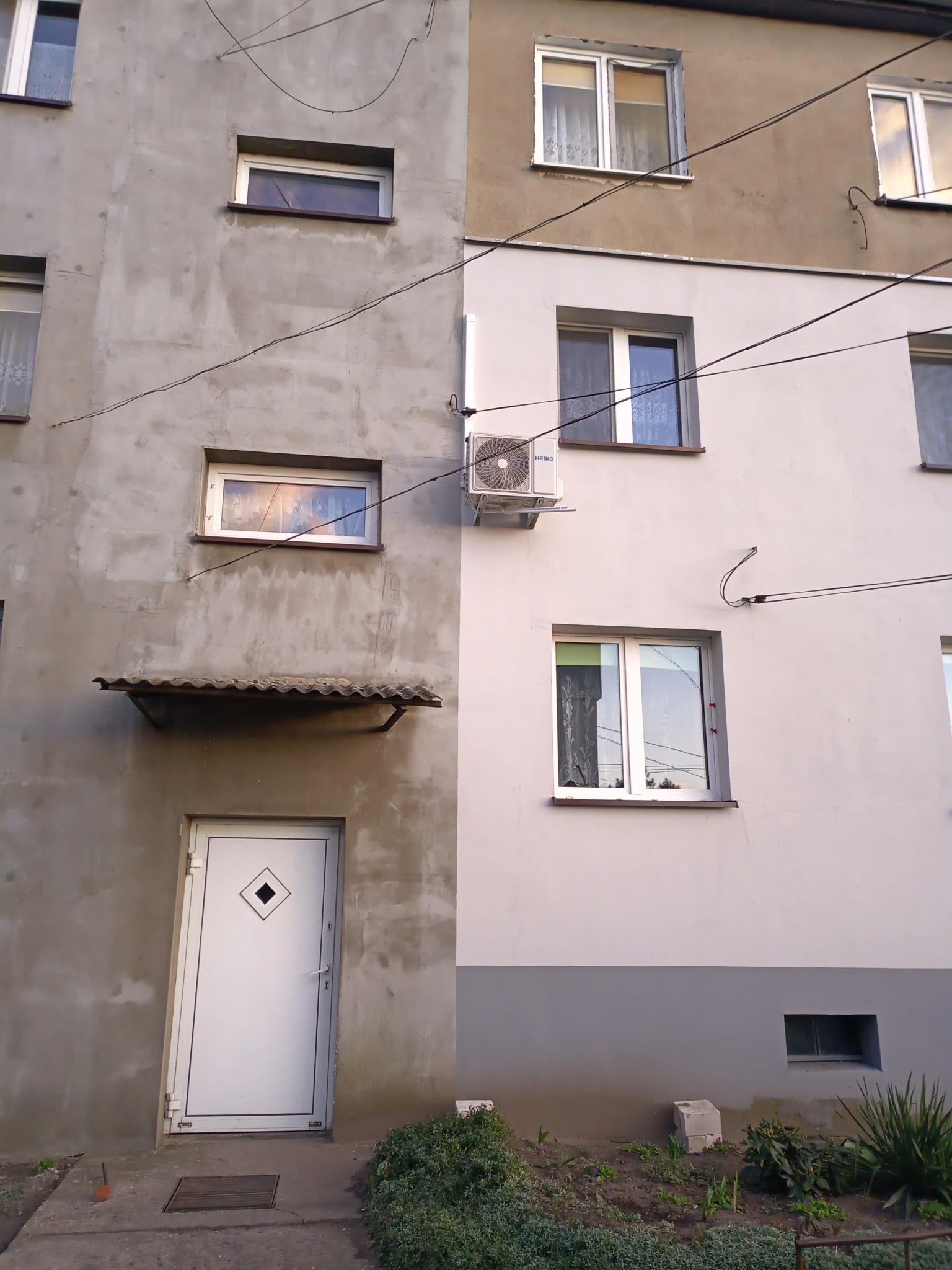 Budynek mieszkalny z zamontowaną jednostką zewnętrzną klimatyzacji, widoczne okna i drzwi wejściowe.