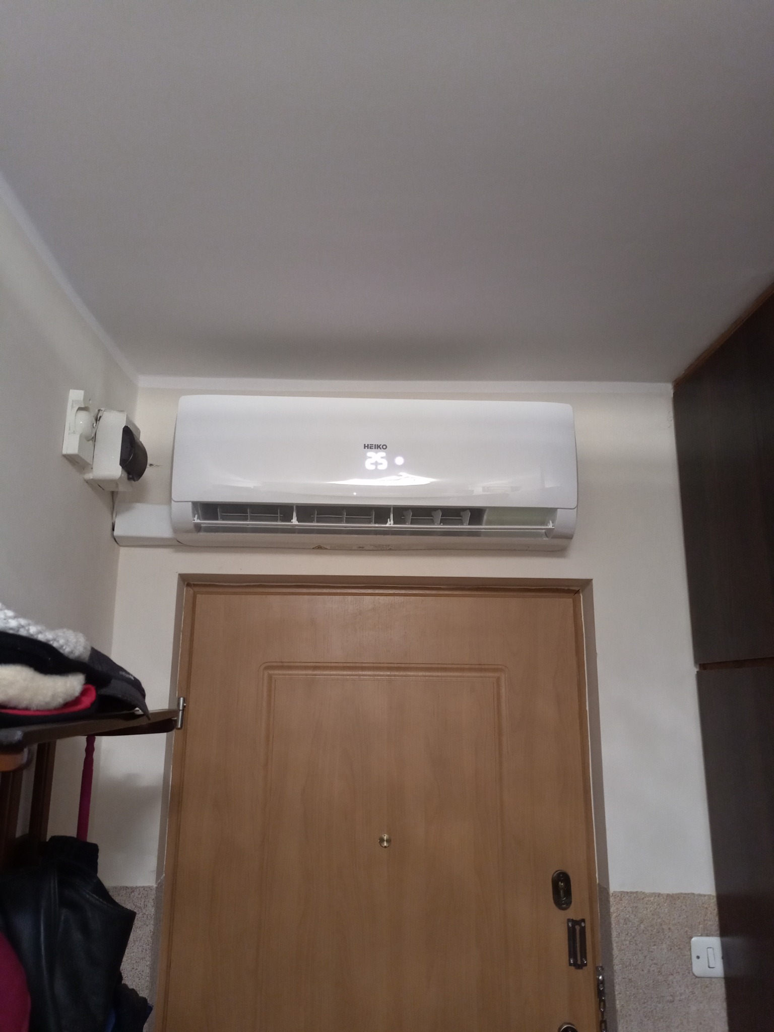 Biały klimatyzator Heiko zamontowany nad drzwiami wewnętrznymi w mieszkaniu, wyświetlający temperaturę 25 stopni Celsjusza, z widocznym gniazdkiem elektrycznym i fragmentem szafy po prawej stronie.