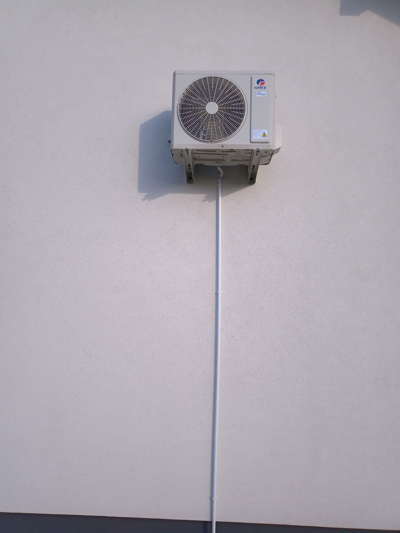 Biała jednostka zewnętrzna klimatyzatora marki Gree zamontowana na jasnej, teksturowanej ścianie budynku, z widoczną rurą odprowadzającą skropliny.