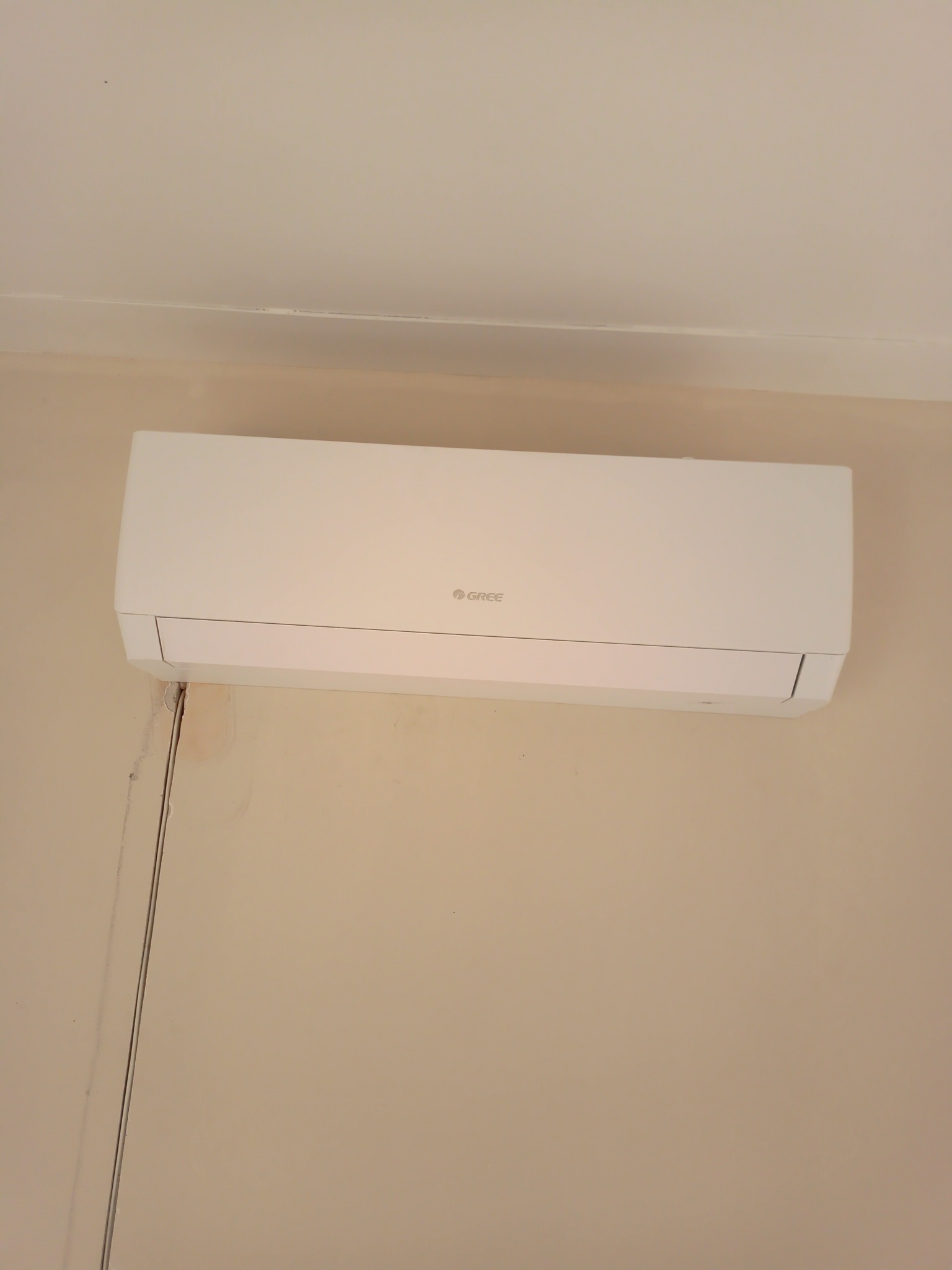 Biały klimatyzator marki Gree zamontowany na beżowej ścianie w pomieszczeniu z widocznymi śladami po instalacji kabli.
