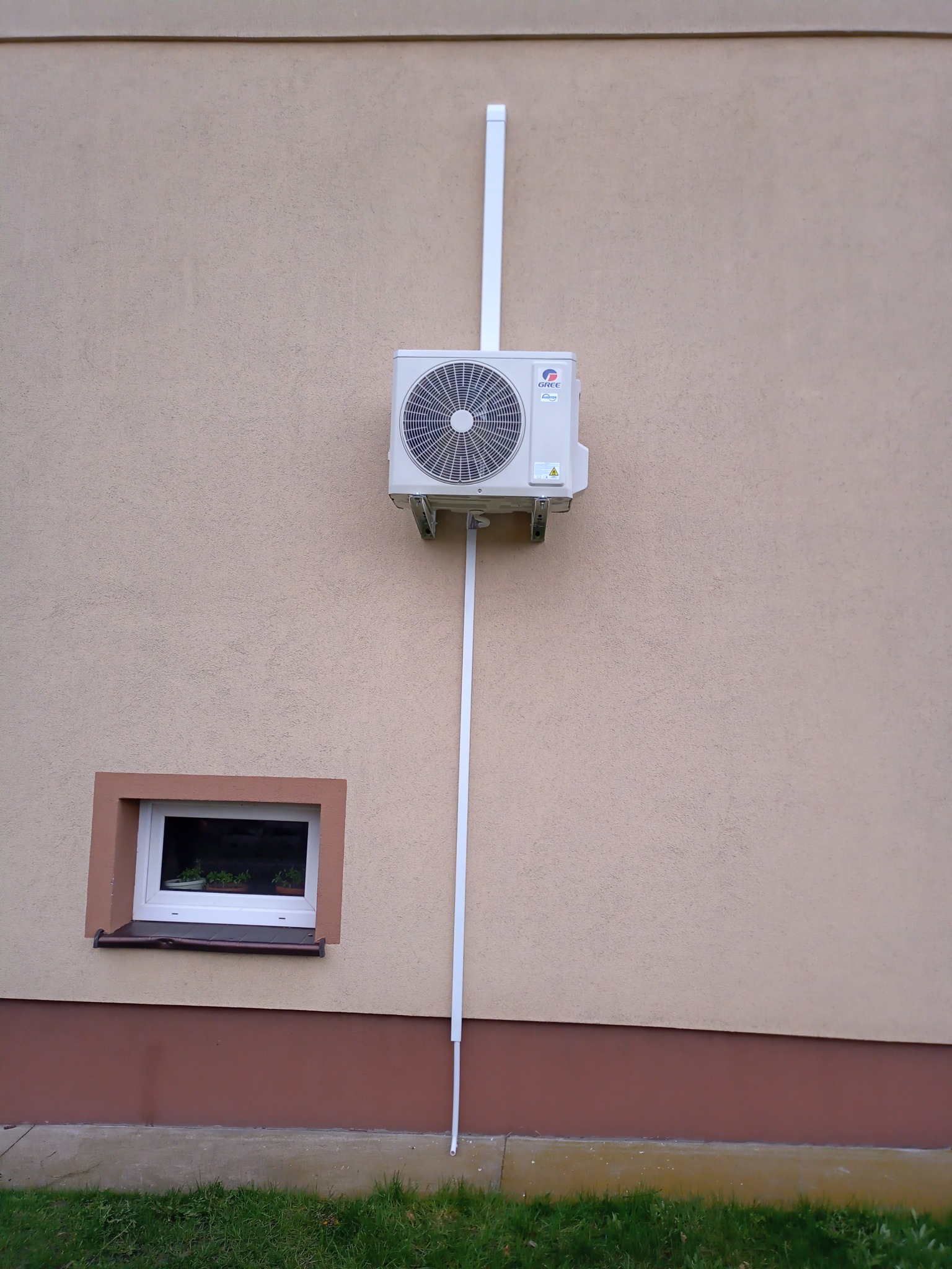 Zewnętrzna jednostka klimatyzacji marki Gree zamontowana na elewacji budynku z widocznymi plastikowymi rurami maskującymi instalację, obok okna z doniczkami.