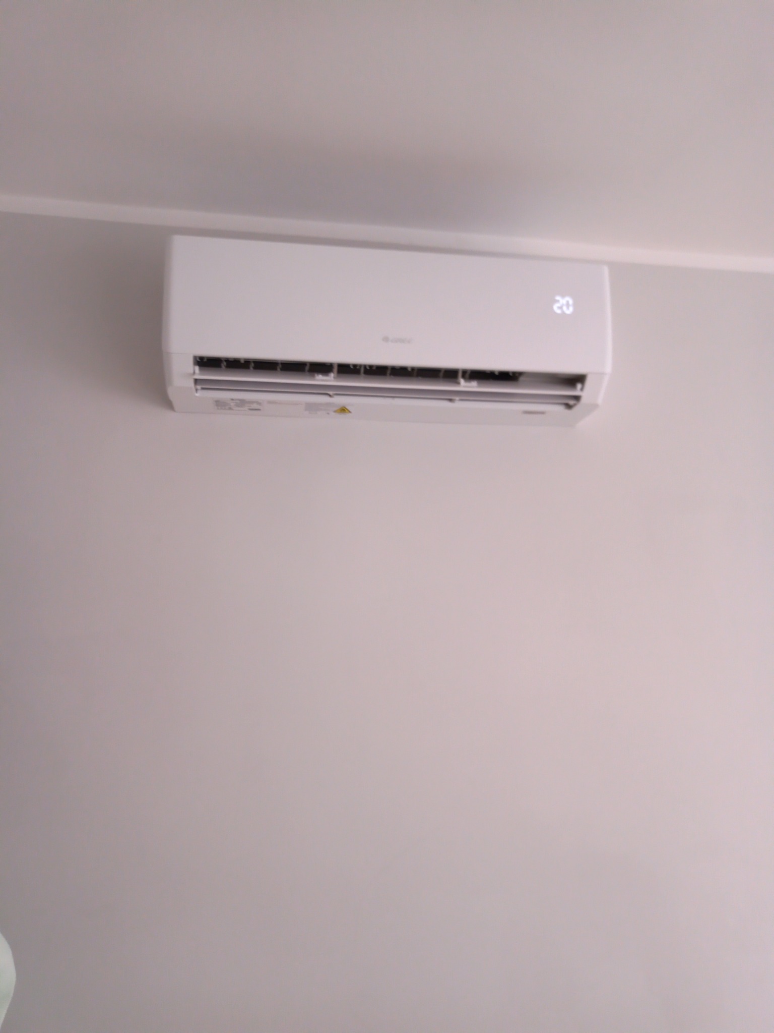 Biała jednostka wewnętrzna klimatyzatora marki Gree, zamontowana na jasnej ścianie, wyświetlająca temperaturę 20 stopni Celsjusza.