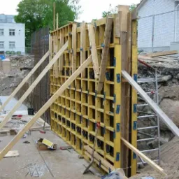 Żółty szalunek ścienny z drewnianymi podporami na placu budowy, widoczna stal zbrojeniowa w tle.