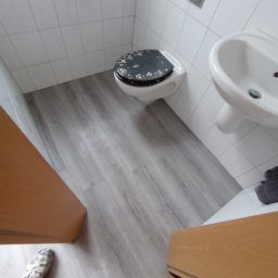 Sucha zabudowa , szpachlowanie , malowanie , podłogi, laminat, winyl - Ukończona łazienka z szarą podłogą imitującą drewno, białe płytki na ścianach, toaleta z czarną deską w kwiaty i biała umywalka.