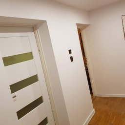Sucha zabudowa , szpachlowanie , malowanie , podłogi, laminat, winyl - Świeżo pomalowane, białe wnętrze z drewnianą podłogą, widoczna nowoczesna biała stolarka drzwiowa z przeszkleniami i włączniki na ścianie.