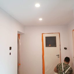 Sucha zabudowa , szpachlowanie , malowanie , podłogi, laminat, winyl - Mężczyzna w ubraniu roboczym maluje ścianę w pomieszczeniu, zabezpieczone drzwi i ościeżnice folią malarską, widoczne dwa wbudowane punkty świetlne w suficie.